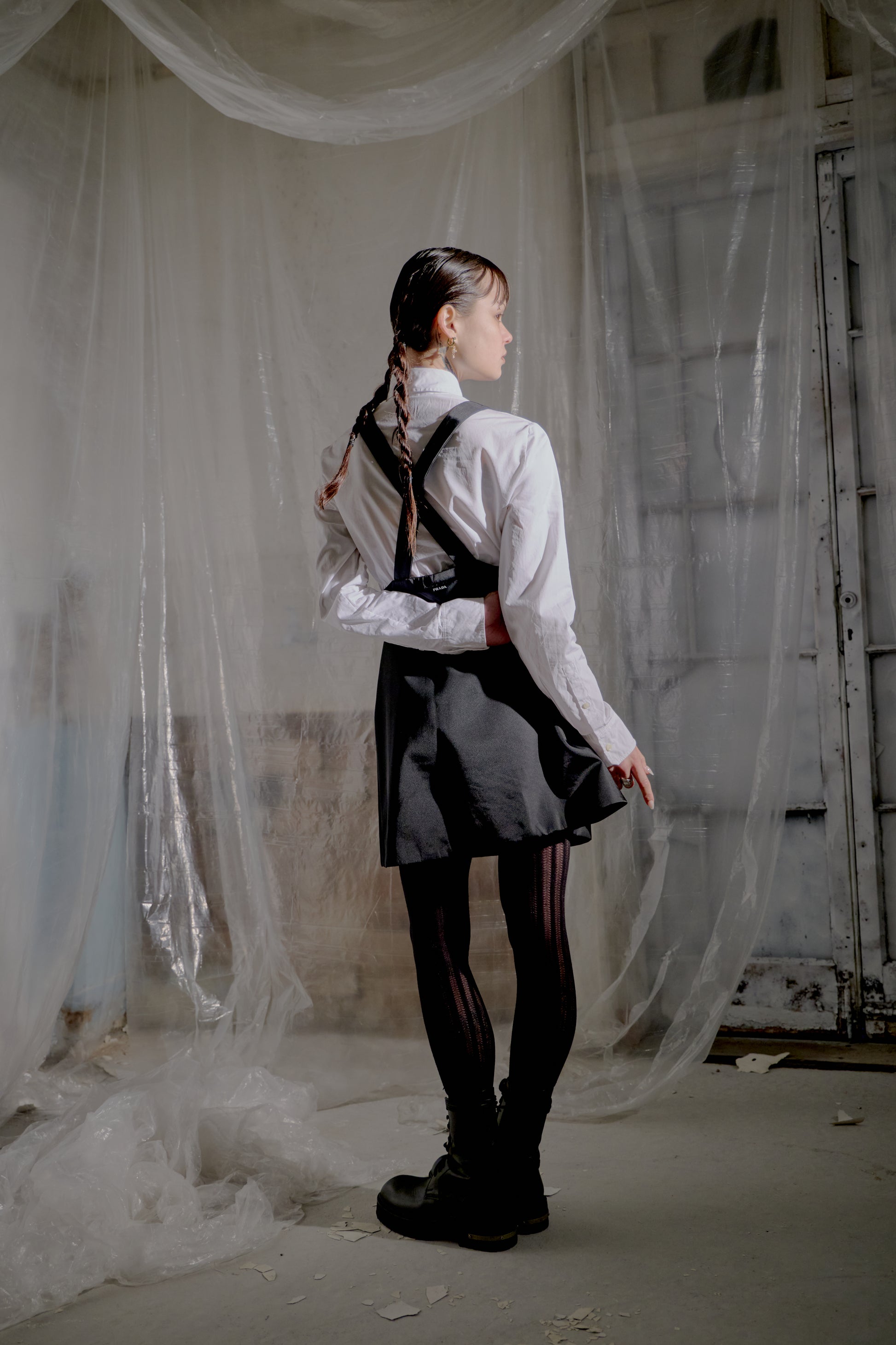 Person wearing a white shirt, black Prada skirt, and suspenders in an abandoned room with curtains.