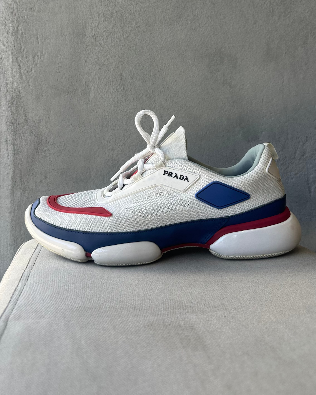 White Prada sneaker with blue and red accents on a gray surface against a gray background