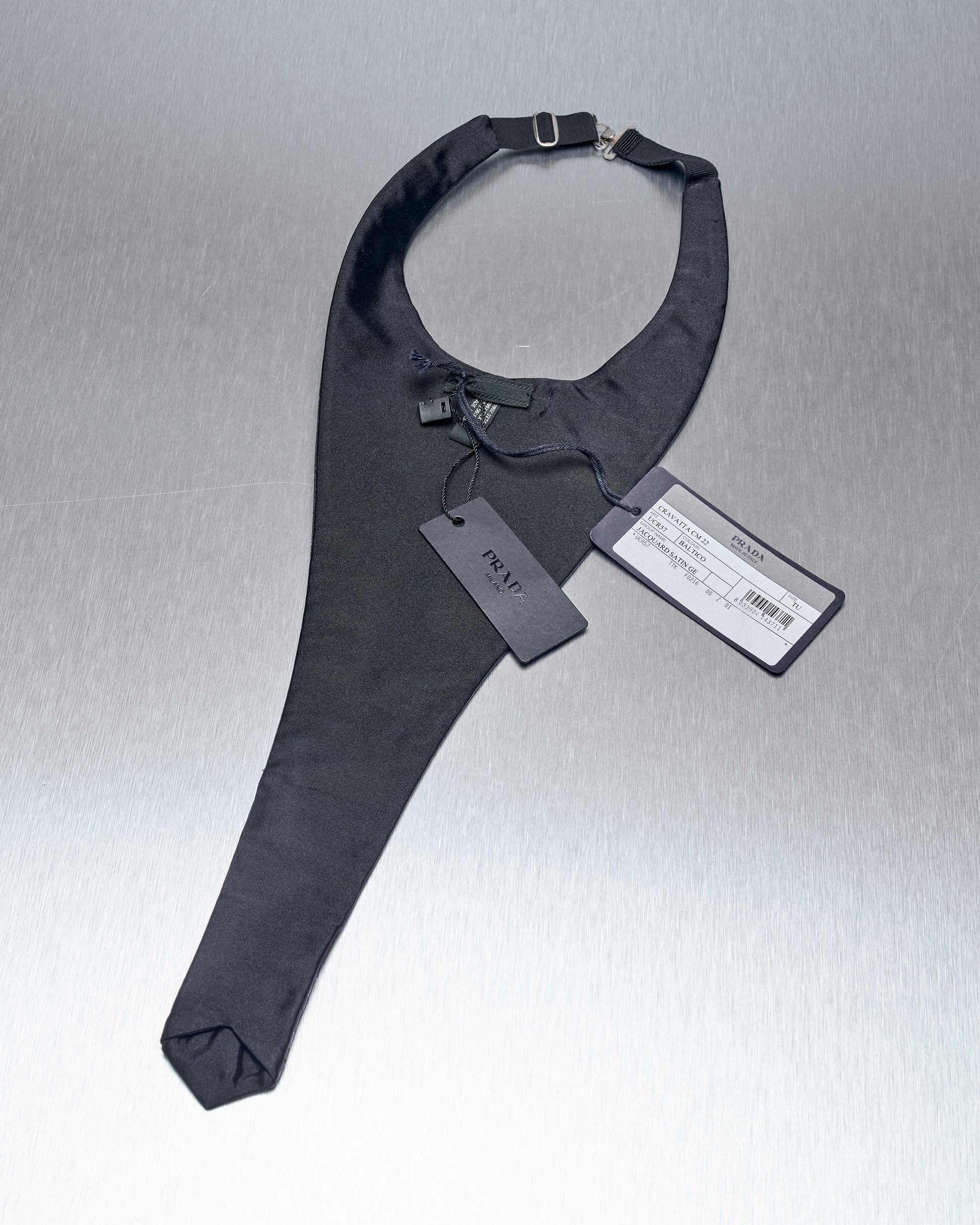 A sleek black Prada cravat neckband with attached tags, lying on a reflective surface. The band is elegant and modern, with a sophisticated tone.