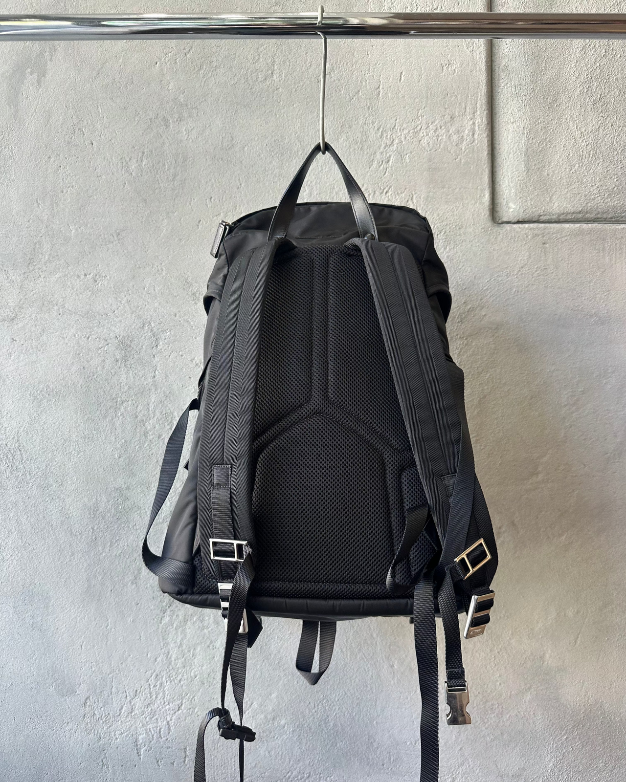 Black Prada backpack with a brand logo hanging on a metal rod against a gray wall.