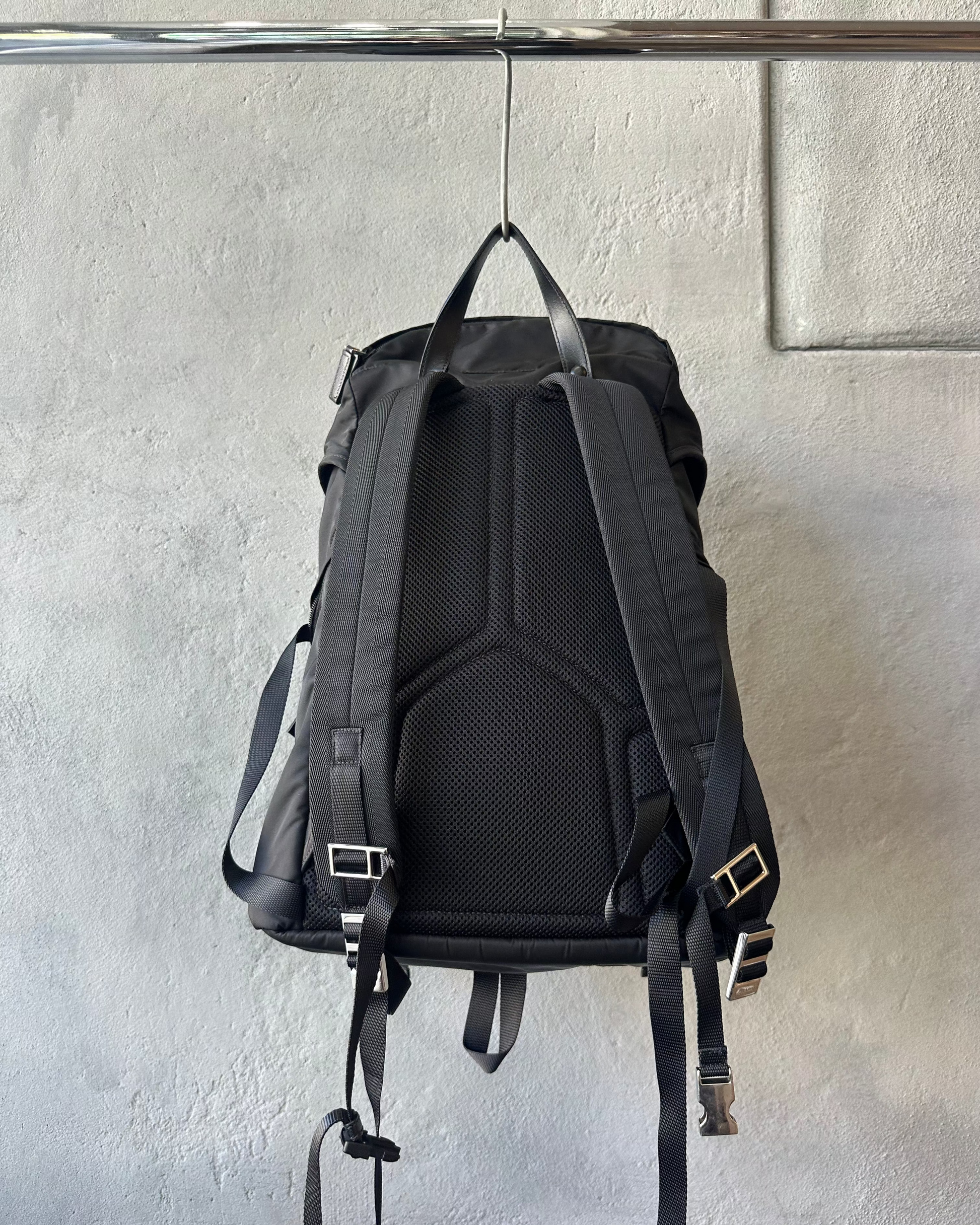 Black Prada backpack hanging on a metal rod against a gray wall.