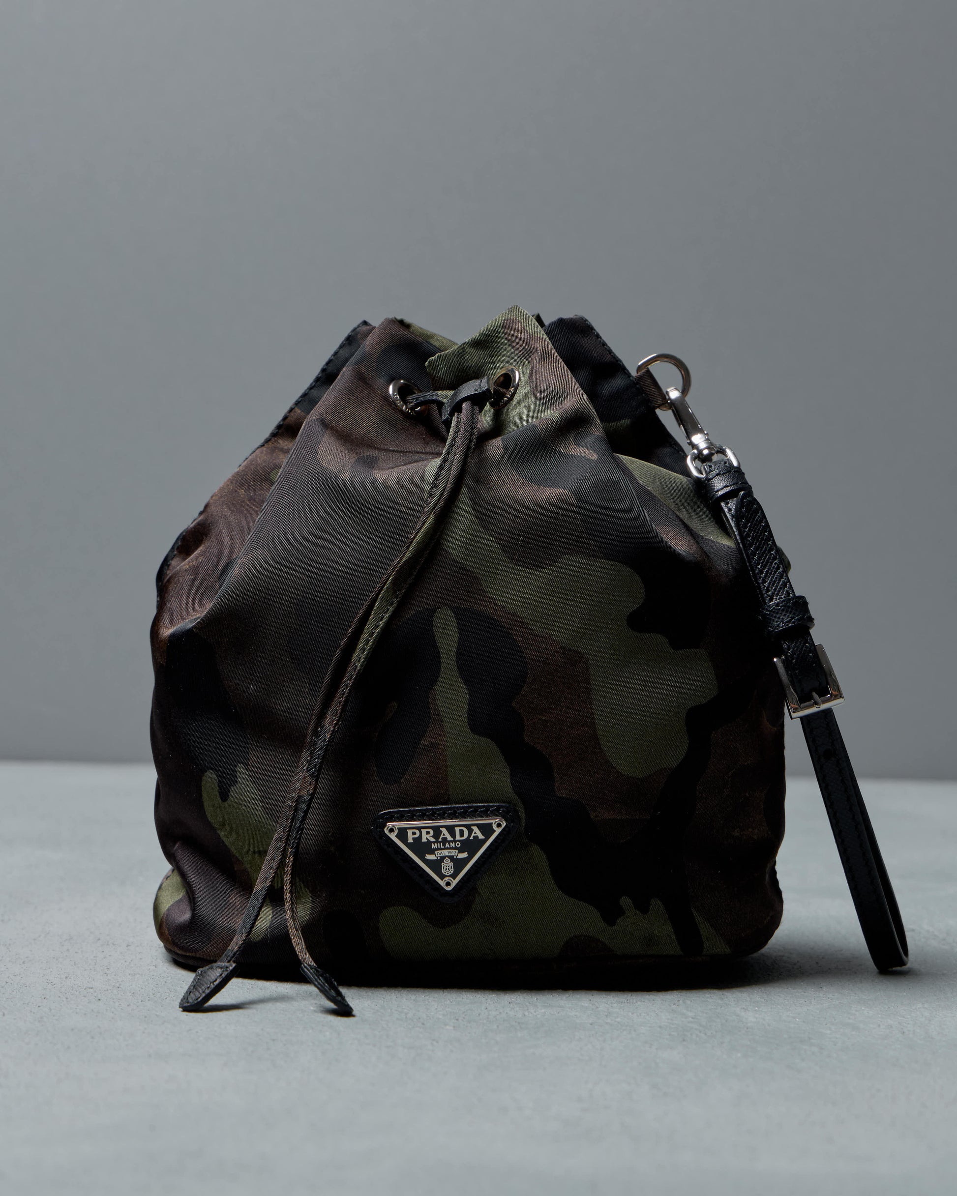Camouflage-patterned Prada bucket bag with a drawstring closure on a gray background, conveying a rugged yet stylish aesthetic.