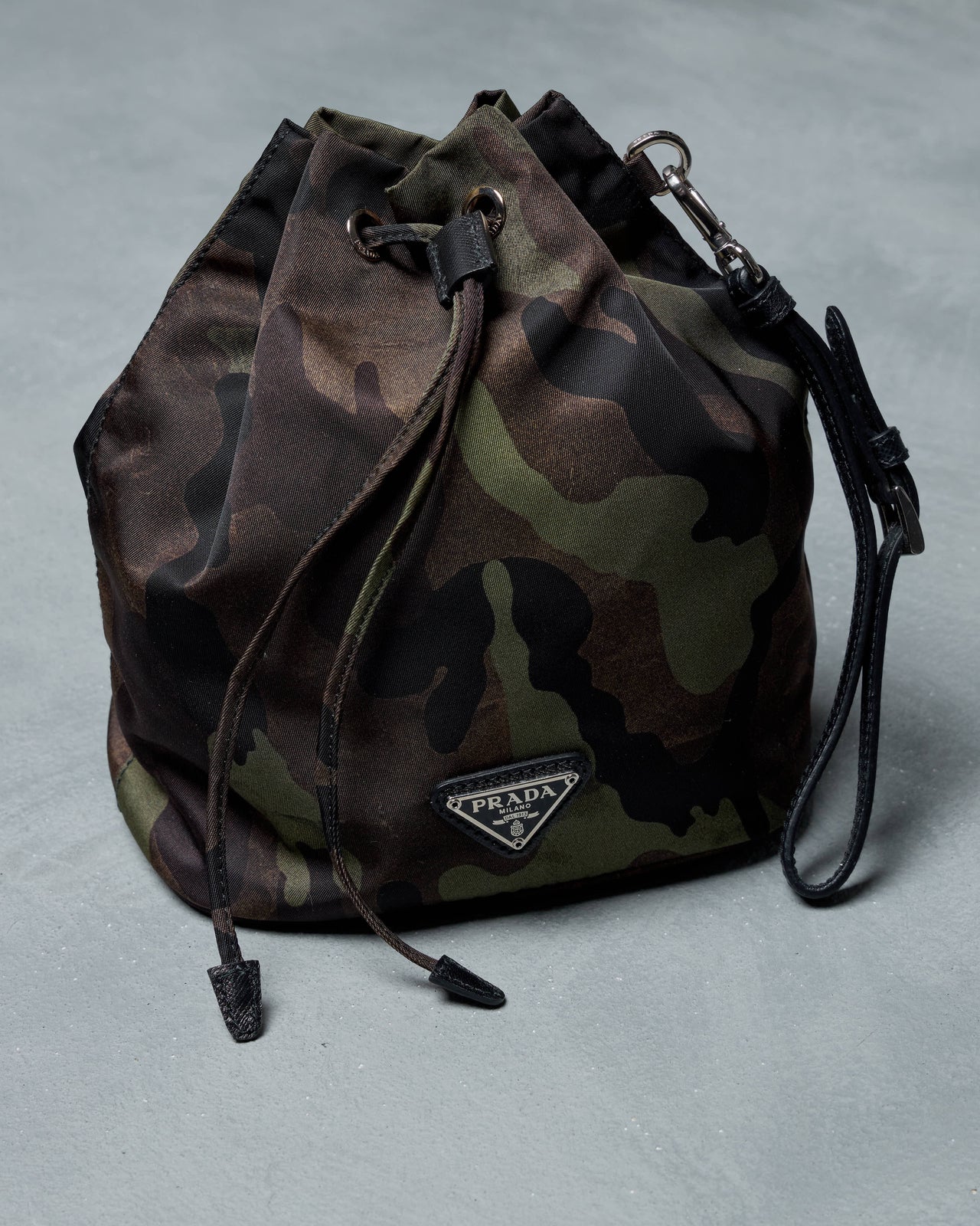 Camouflage-patterned Prada bucket bag with a drawstring closure on a gray background, conveying a rugged yet stylish aesthetic.