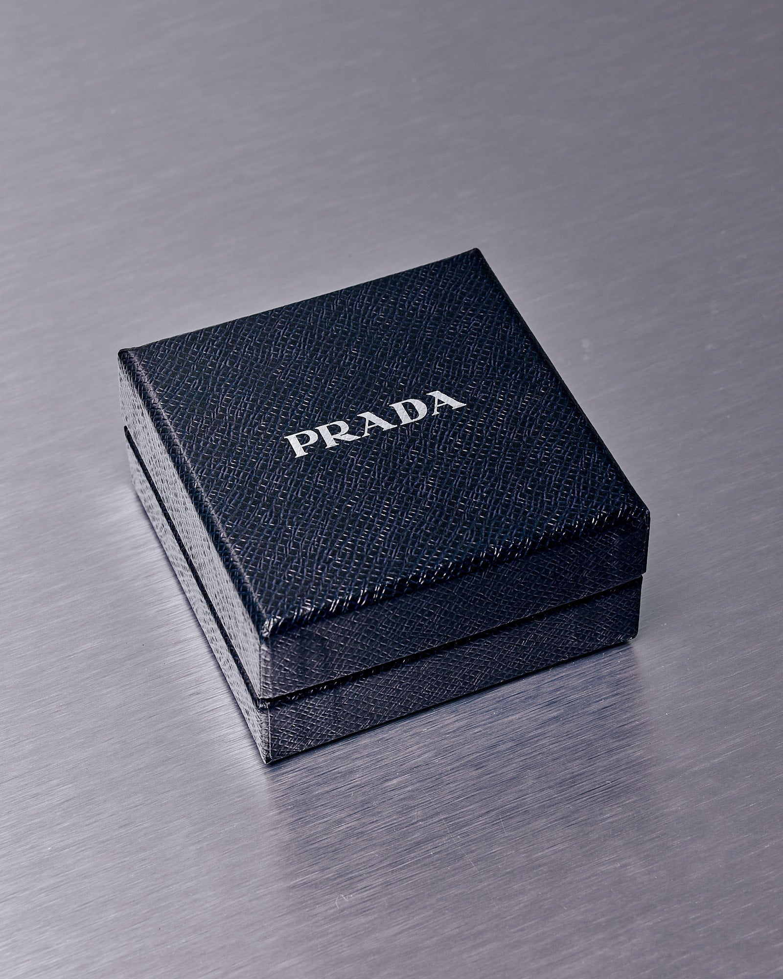 A textured black Prada box on a metallic surface, exuding luxury and elegance. The white Prada logo is prominently displayed on the lid.