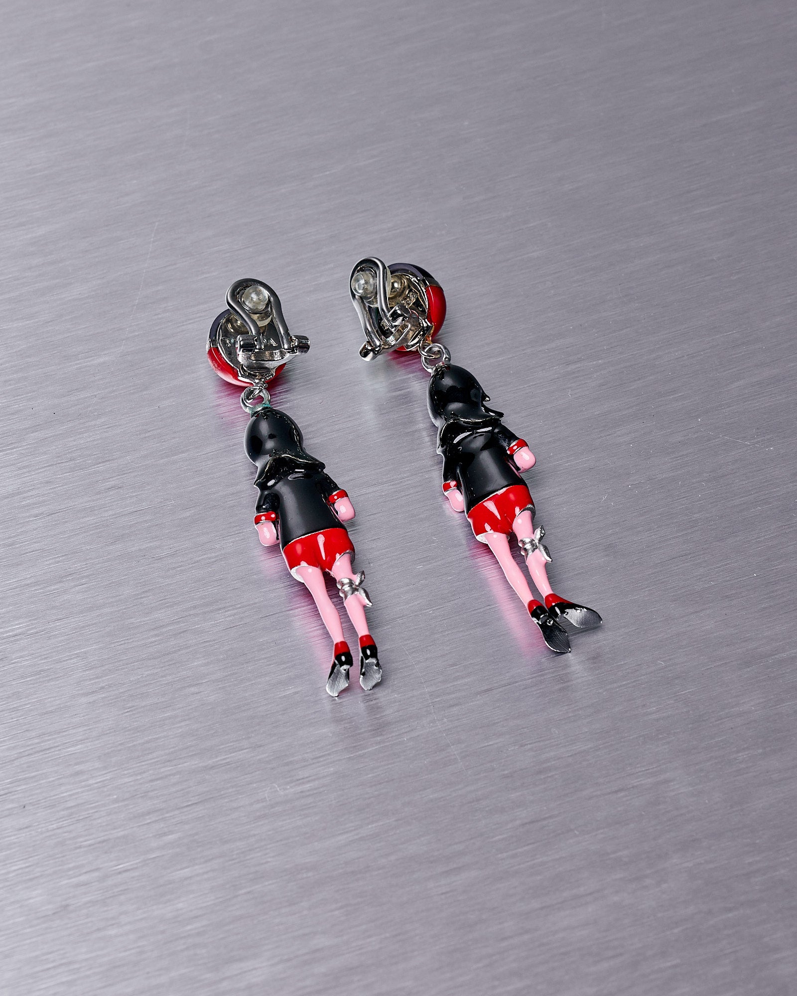 Prada earrings with red skirts, black tops, and silver shoes set on a brushed metal surface. 