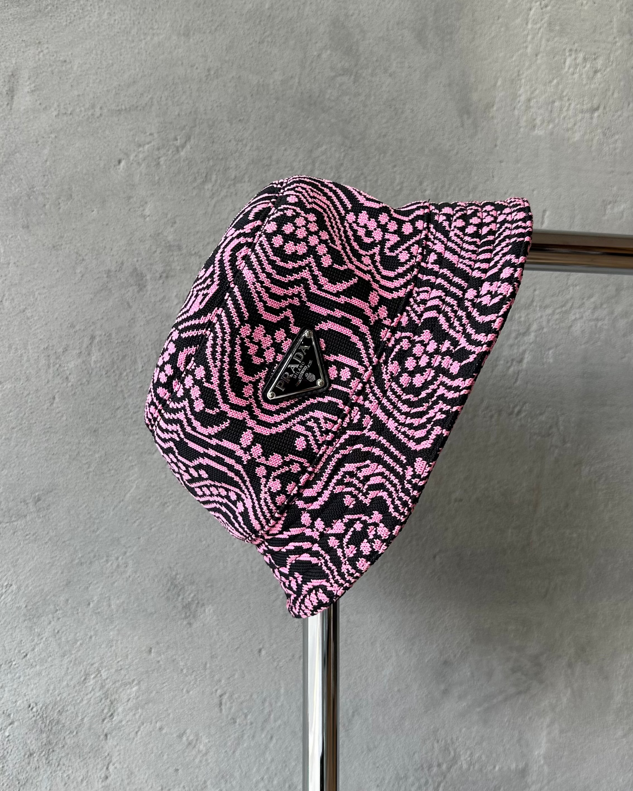 Pink and black patterned Prada bucket hat on a metal rod against a gray wall