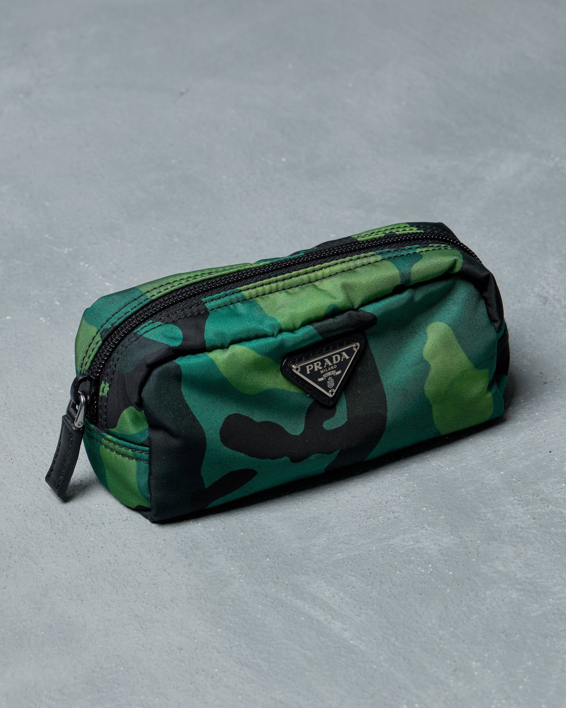 Green camouflage Prada pouch with black accents on a gray textured surface, featuring a prominent "Prada" logo on the front.
