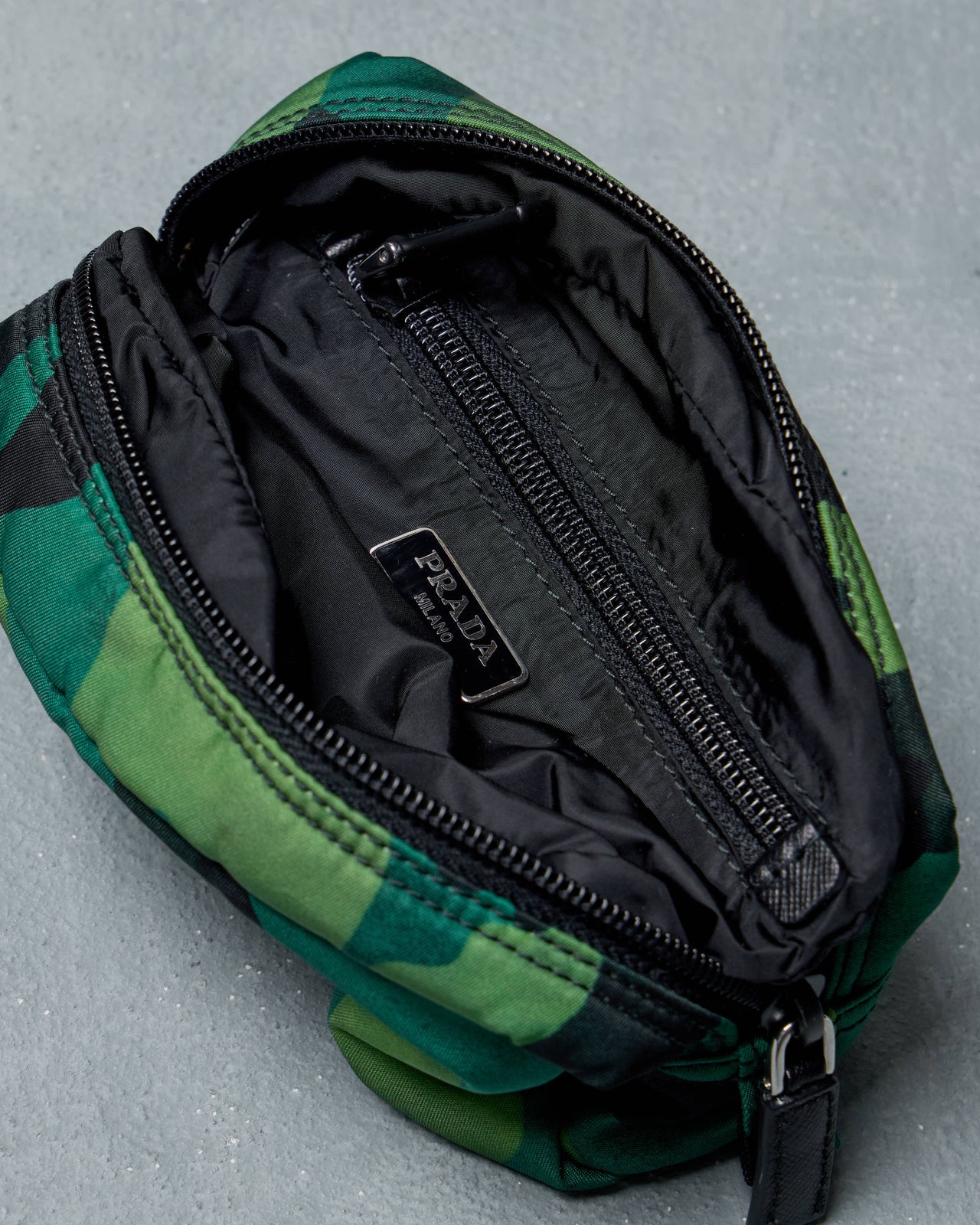 Green camouflage Prada pouch with black accents on a gray textured surface, featuring a prominent "Prada" logo on the front.