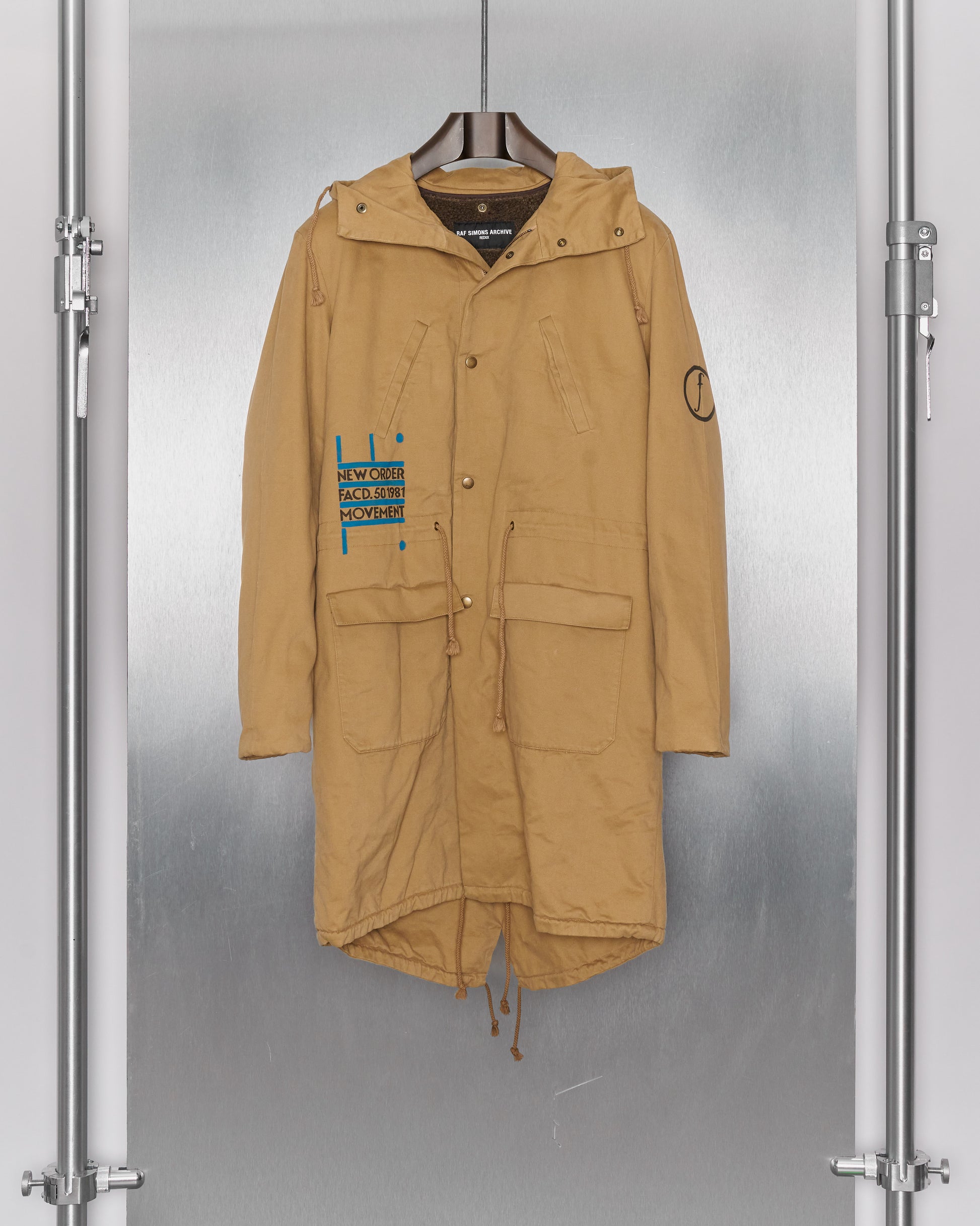 Tan hooded Raf Simons coat with large front pockets and buttoned closure, hanging on a hanger. Left chest has blue text and graphics; right sleeve has a circle design.