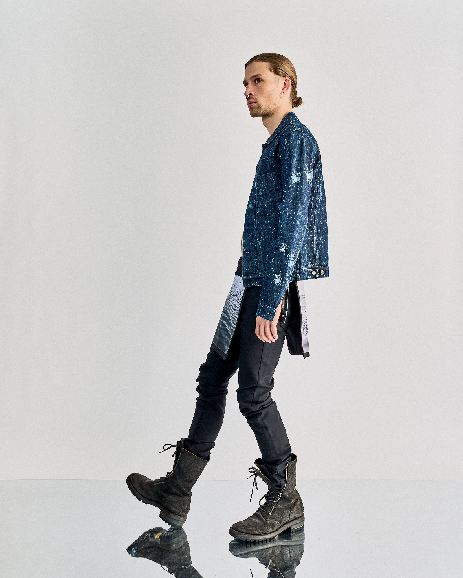 A person in profile wearing a blue denim jacket, black Raf Simons pants, and rugged boots walks confidently on a reflective surface.