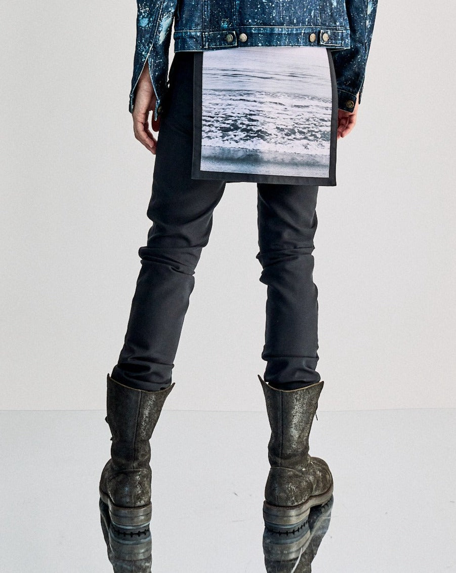Back view of a person wearing a denim jacket with seascape print Raf Simons pants dangling from it, dark pants, and rugged boots, standing on a reflective surface.