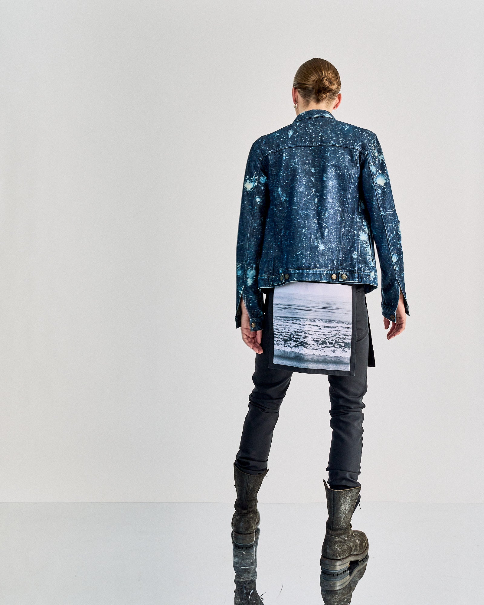 Back view of a person wearing a denim jacket with seascape print Raf Simons pants dangling from it, dark pants, and rugged boots, standing on a reflective surface.