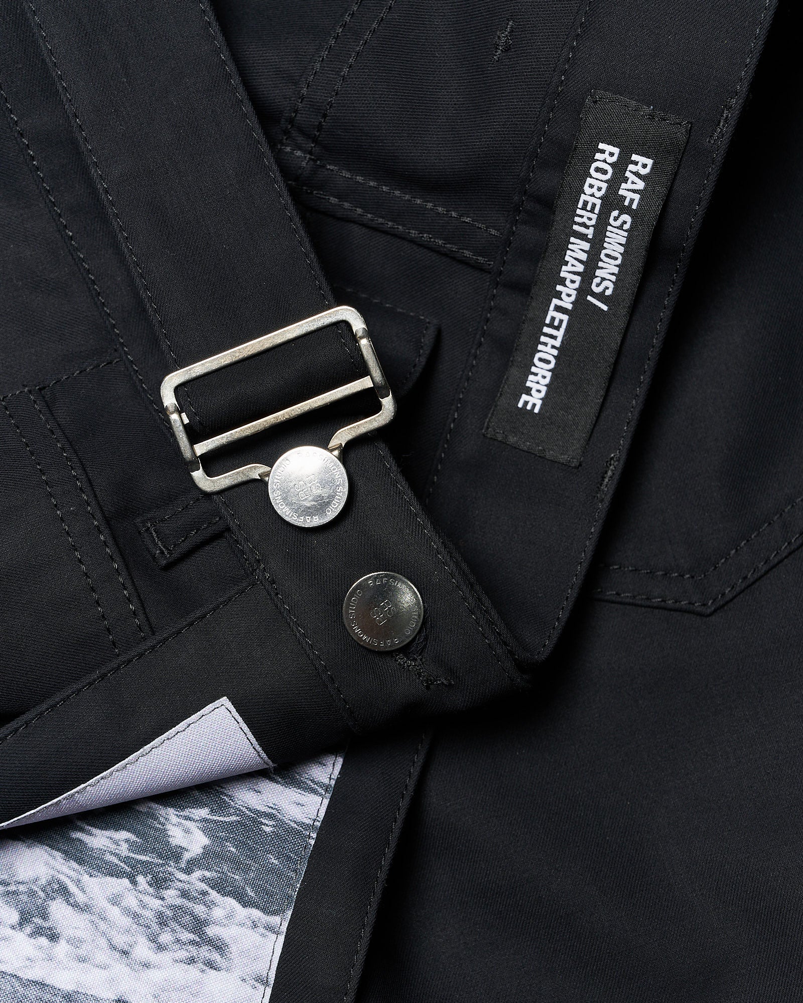 Close-up of a black garment featuring a metal buckle, silver buttons, and a stitched label with "RAF SIMONS / ROBERT MAPPLETHORPE." A hint of a textured fabric peeks from underneath, adding contrast.