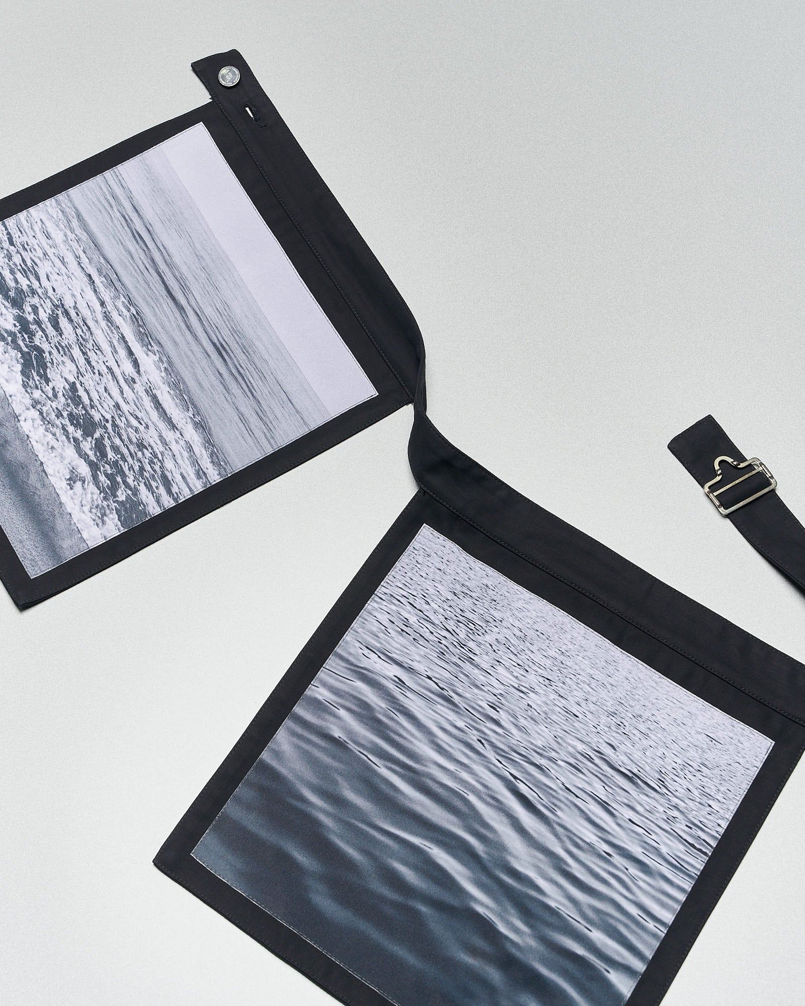 Two black fabric panels from Raf Simons featuring black-and-white ocean photographs are laid flat. One panel shows waves, the other calm waters, creating a serene mood.