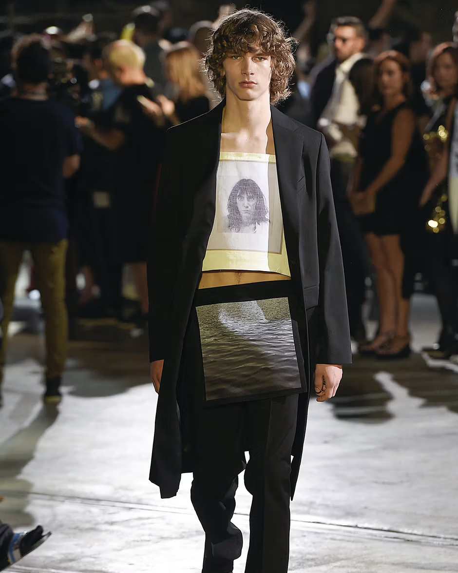Model walking on a Raf Simons runway wearing a black coat and graphic t-shirt with a portrait.