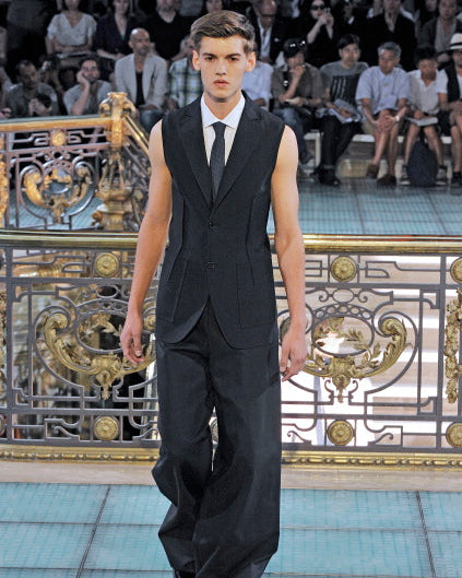A male model wearing a black long-sleeved Raf Simons blazer with a notch lapel and two button closures, featuring external darting on the chest and back of the lower waist, with two patch pockets and an internal pocket.