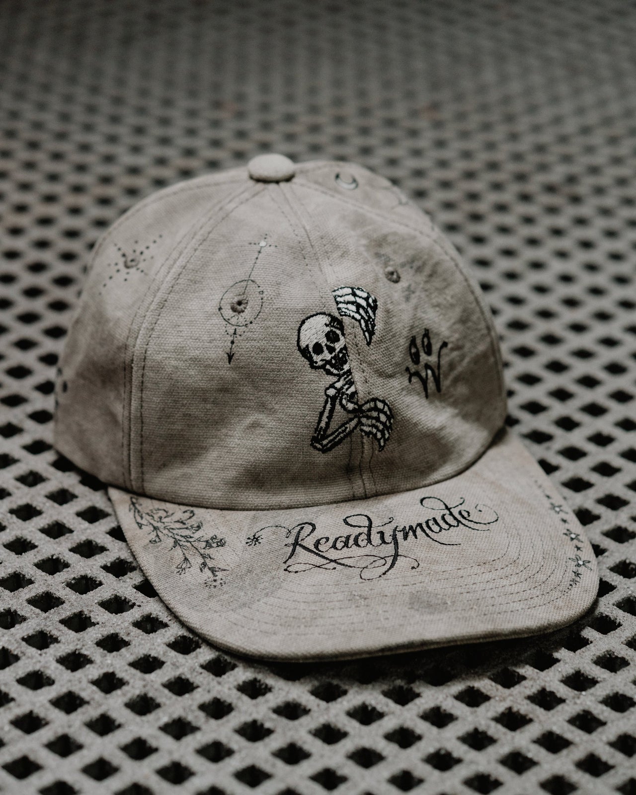 A beige distressed baseball Readymade x Dr Woo cap with a graphic of a skull and crossbones on the front and the brand name 'Readymade' written in a cursive script.