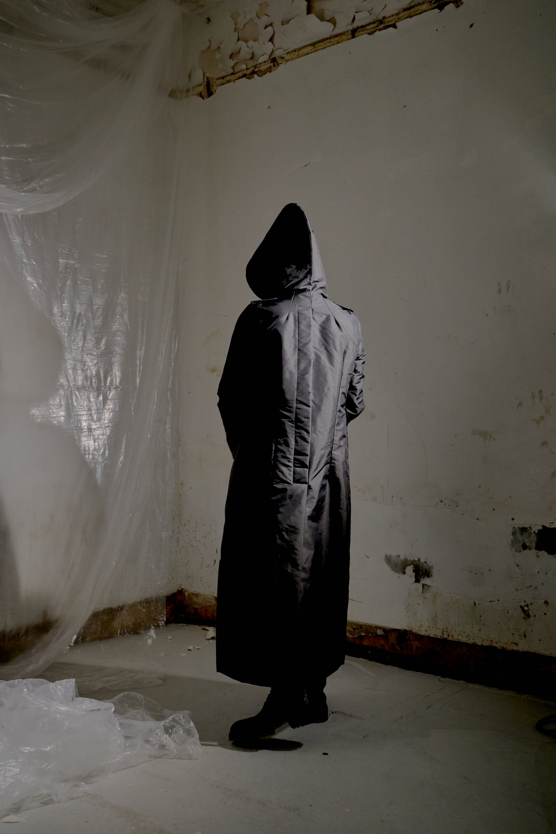 Person wearing a long Rick Owens coat in a dilapidated room with white plastic sheeting.