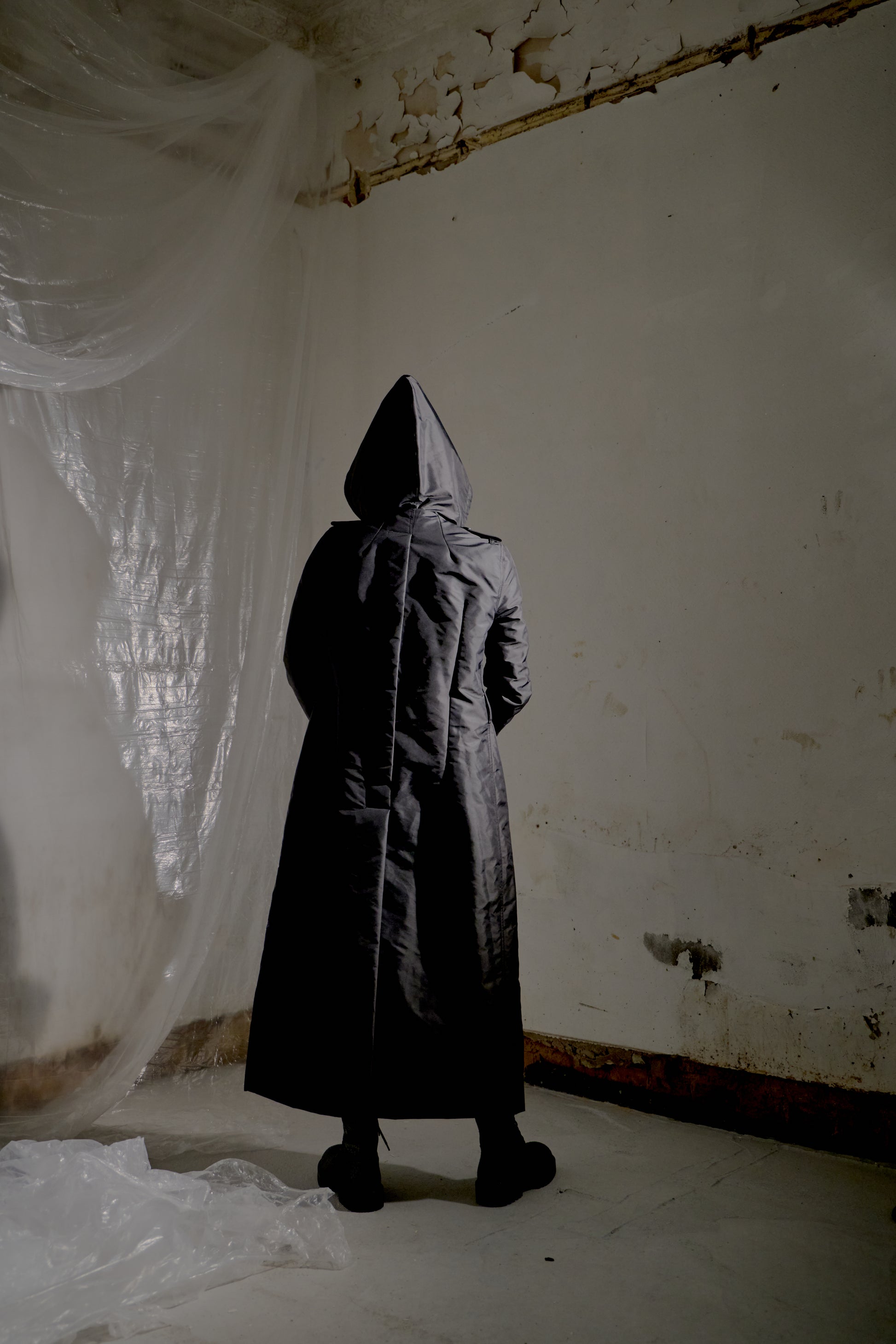Person wearing a long black Rick Owens coat and hood in a dilapidated room with white plastic sheeting.