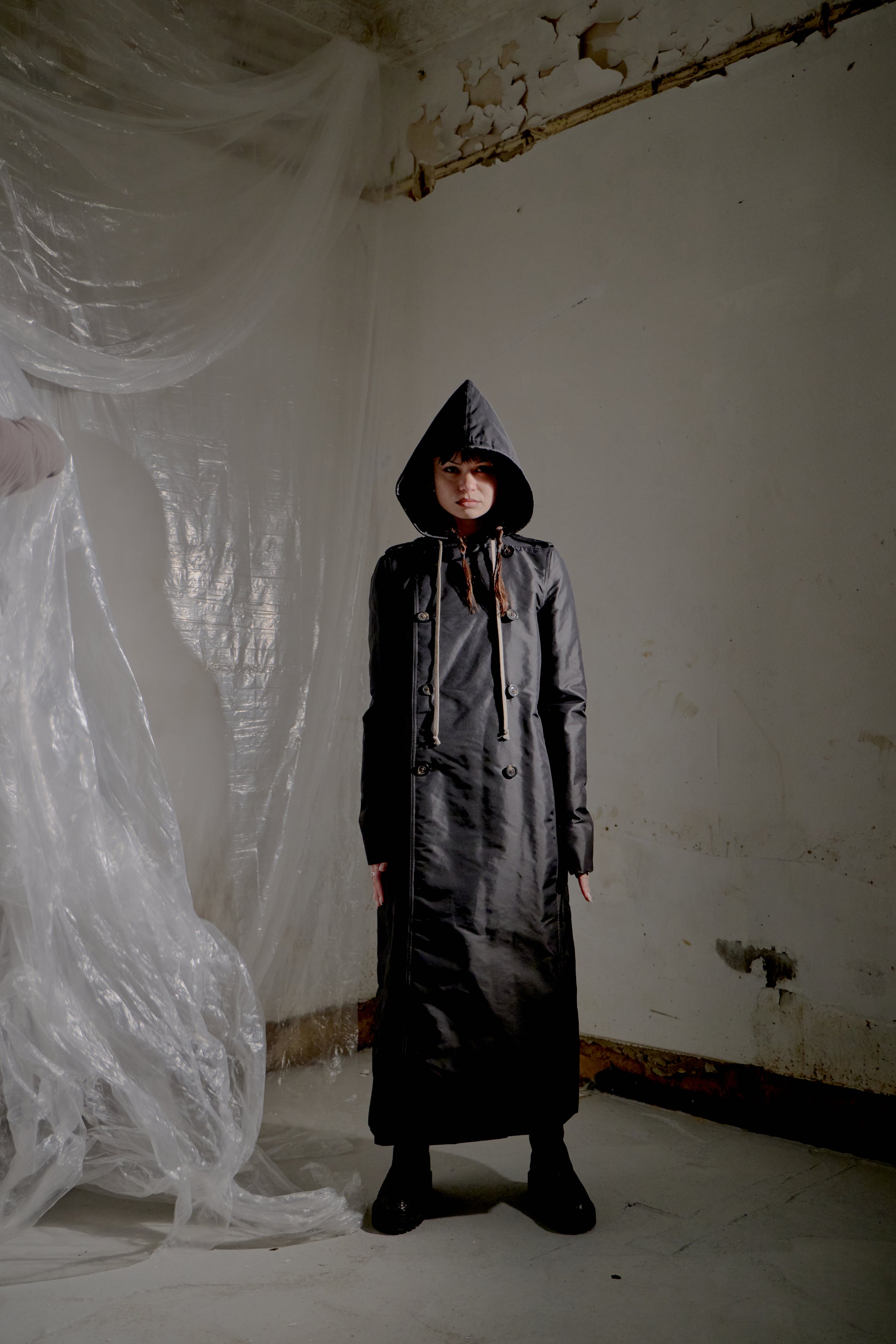 Person wearing a long black Rick Owens coat in a room with white walls and draped fabric.
