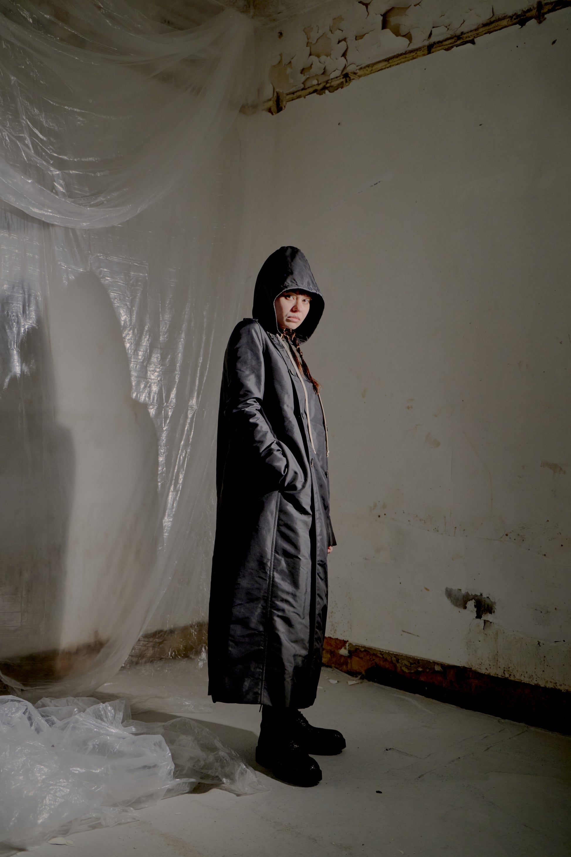 Person wearing a long Rick Owens coat in a room with white walls and a plastic sheet.