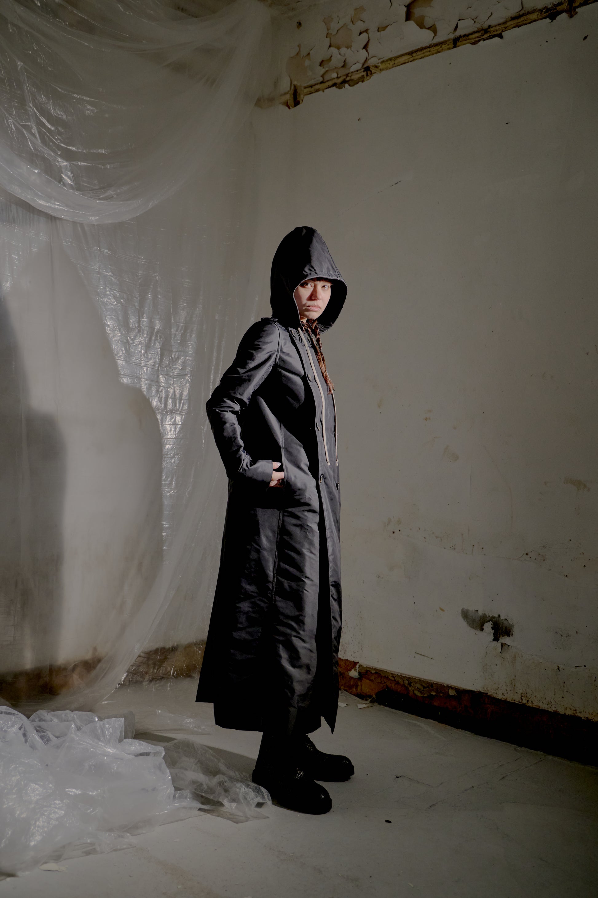 Person wearing a long black Rick Owens coat in a dilapidated room with white walls and plastic sheeting.