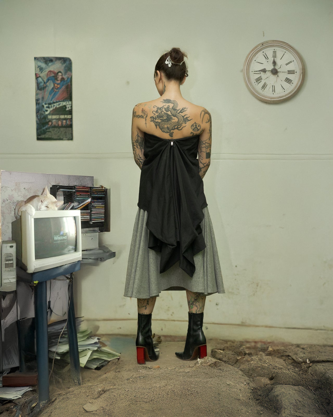 A person standing in a room with a computer monitor, wearing a Rick Owens bustier, a knee-length skirt, and boots. The room has a clock on the wall and appears to be cluttered.