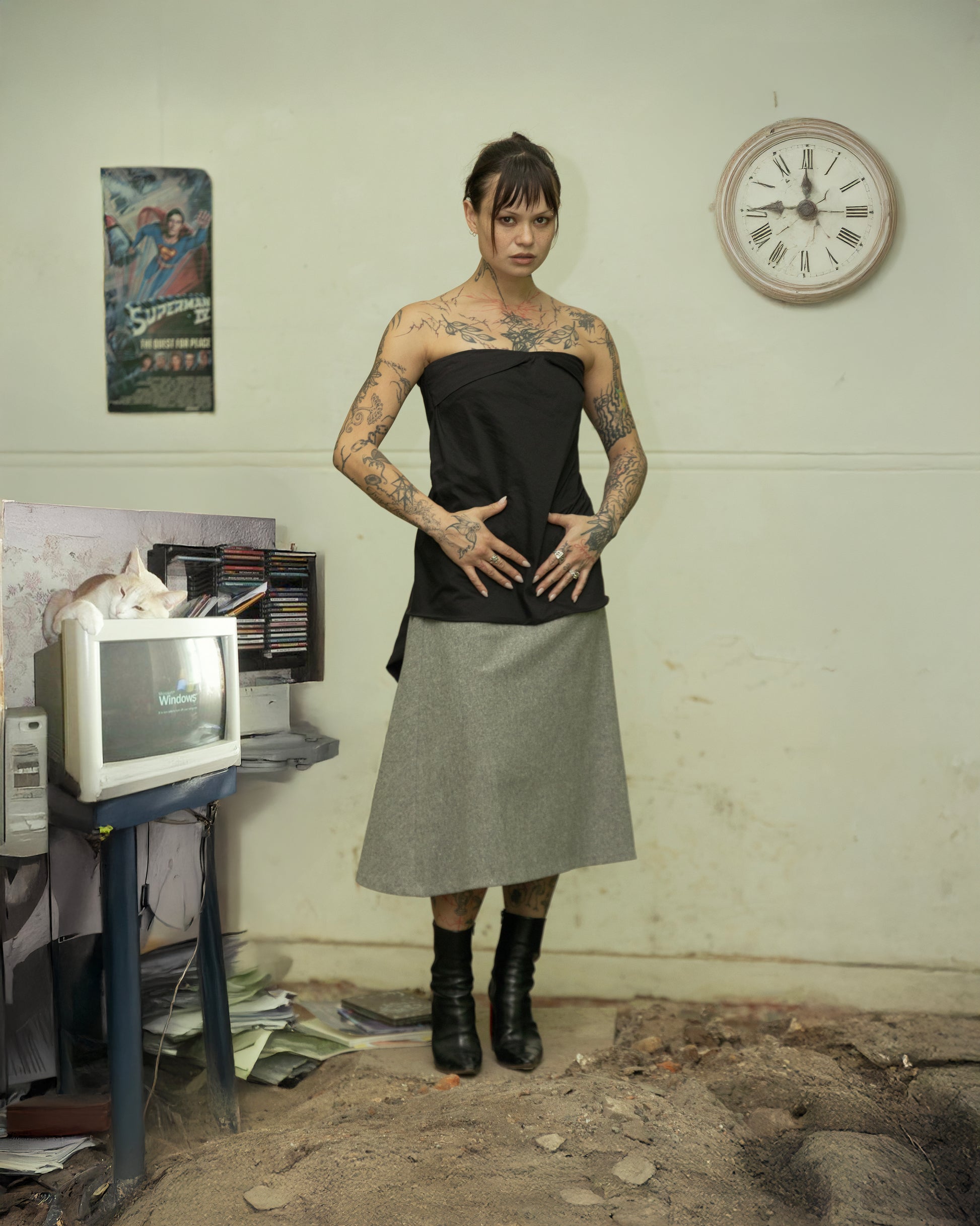 Person wearing a black Rick Owens top and gray skirt standing in a room with a clock on the wall.