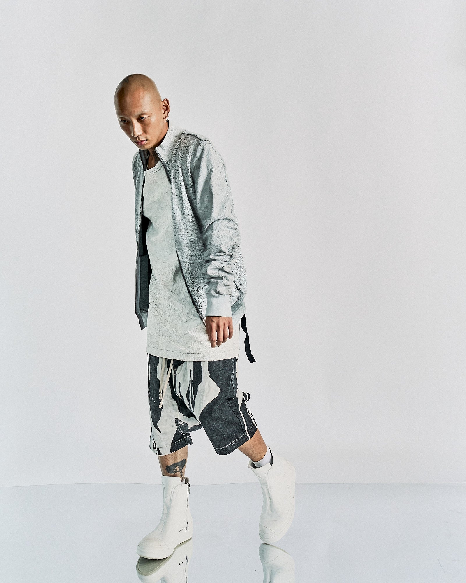 Person in a minimalist, edgy Rick Owens outfit featuring a light gray textured jacket, long white shirt, abstract print shorts, and white boots. Calm expression.
