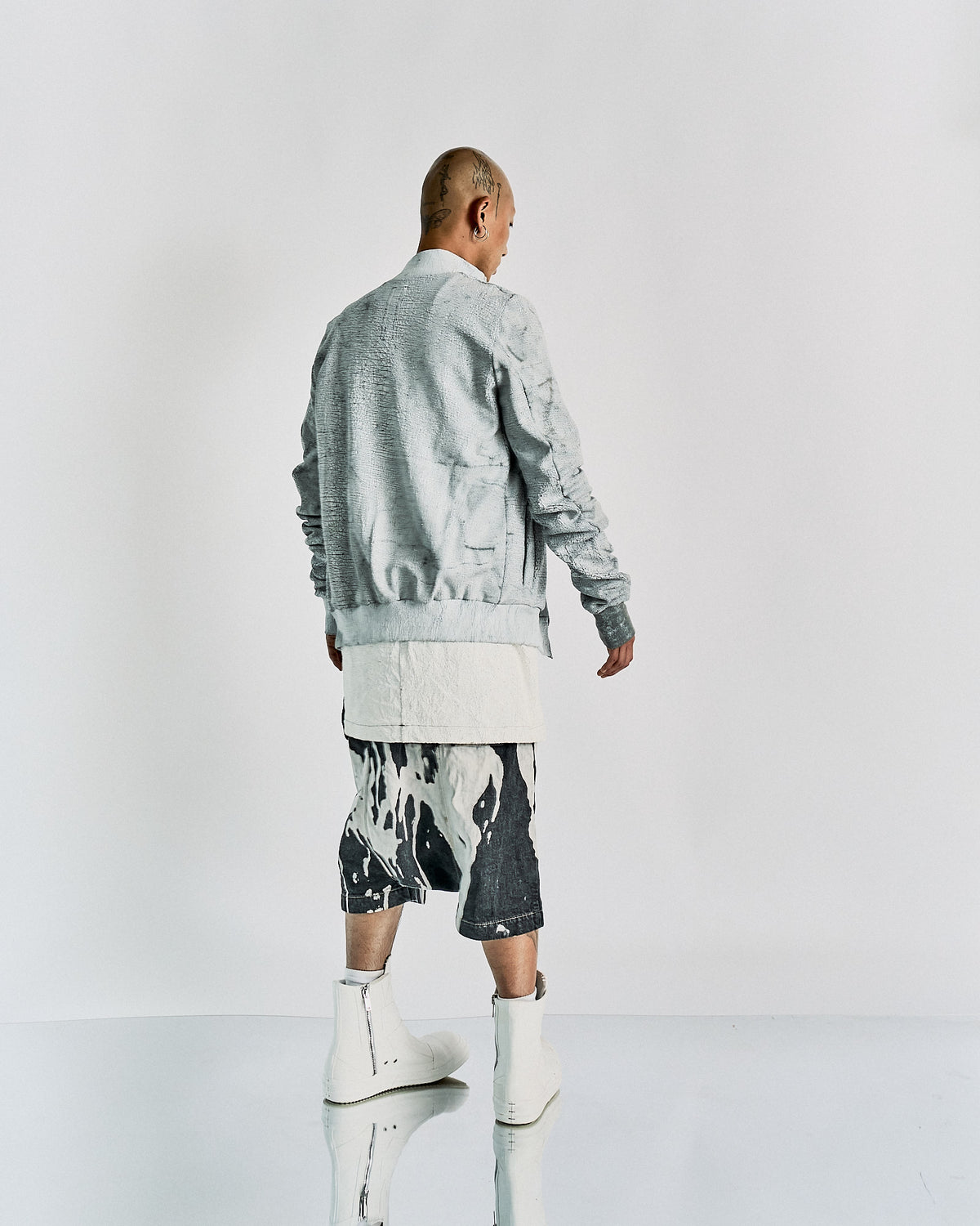 Person in a minimalist, edgy Rick Owens outfit featuring a light gray textured jacket, long white shirt, abstract print shorts, and white boots. Calm expression.