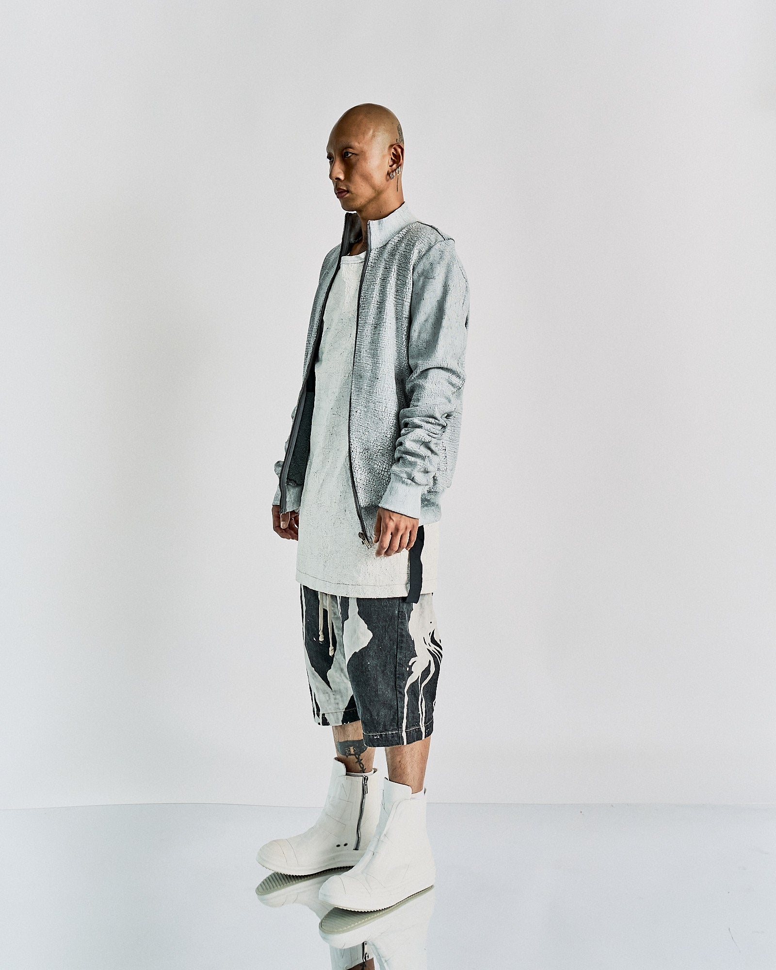 A person stands in profile, wearing a light gray Rick Owens jacket, long white shirt, abstract black and white shorts, and white high boots. The tone is modern and edgy.