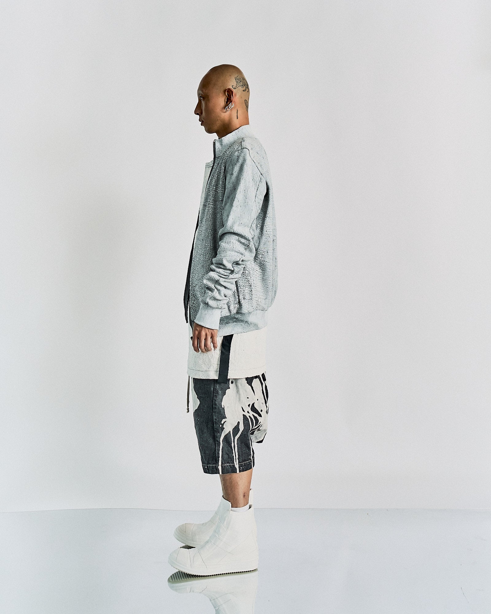 A person stands in profile, wearing a light gray Rick Owens jacket, long white shirt, abstract black and white shorts, and white high boots. The tone is modern and edgy.