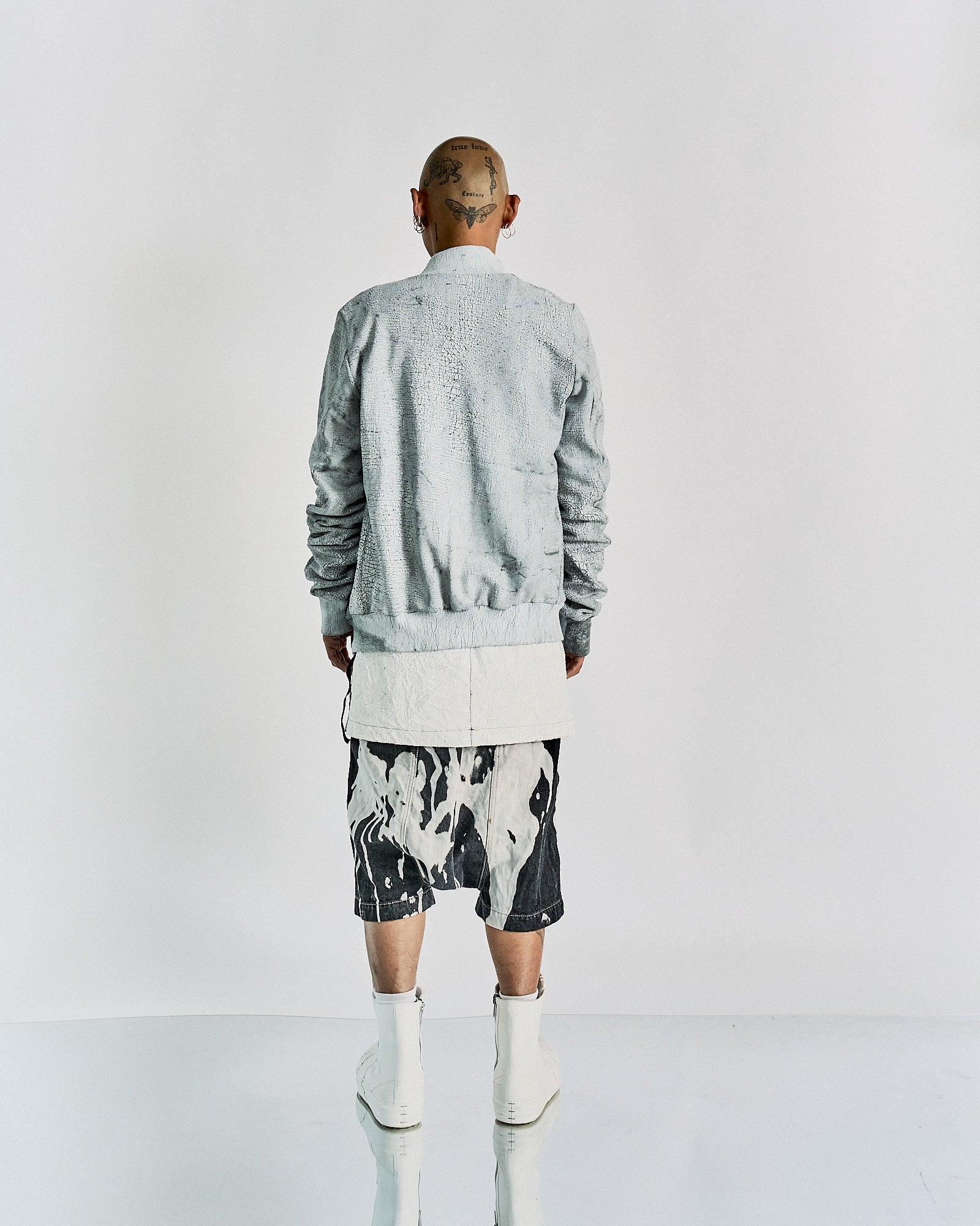 A person with tattoos on their shaved head is wearing a textured gray Rick Owens jacket, patterned shorts, and white boots. The tone is modern and edgy.