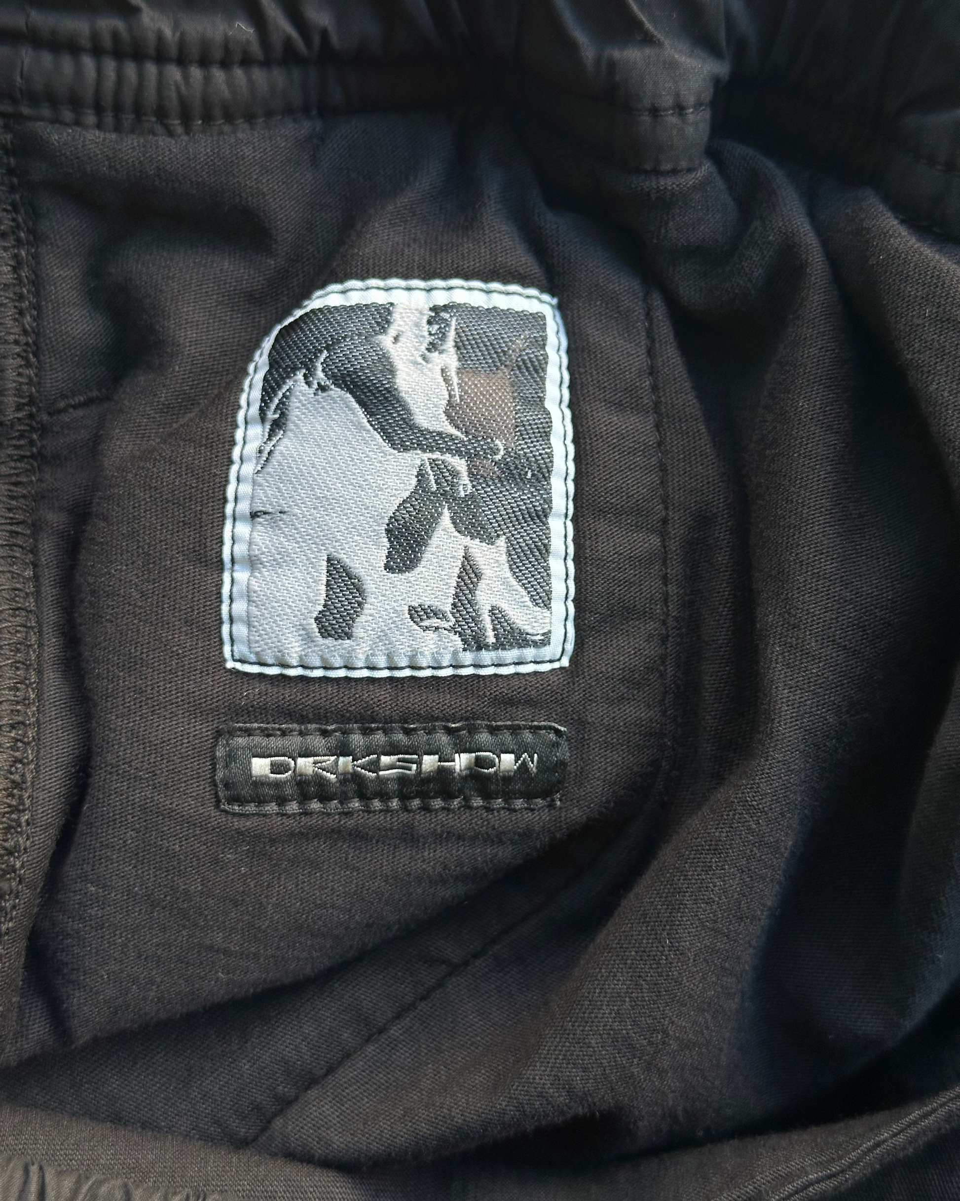 Black fabric from a pair of Rick Owens shorts with a logo patch featuring a person on a horse, brand 'DRKSHDW' visible.