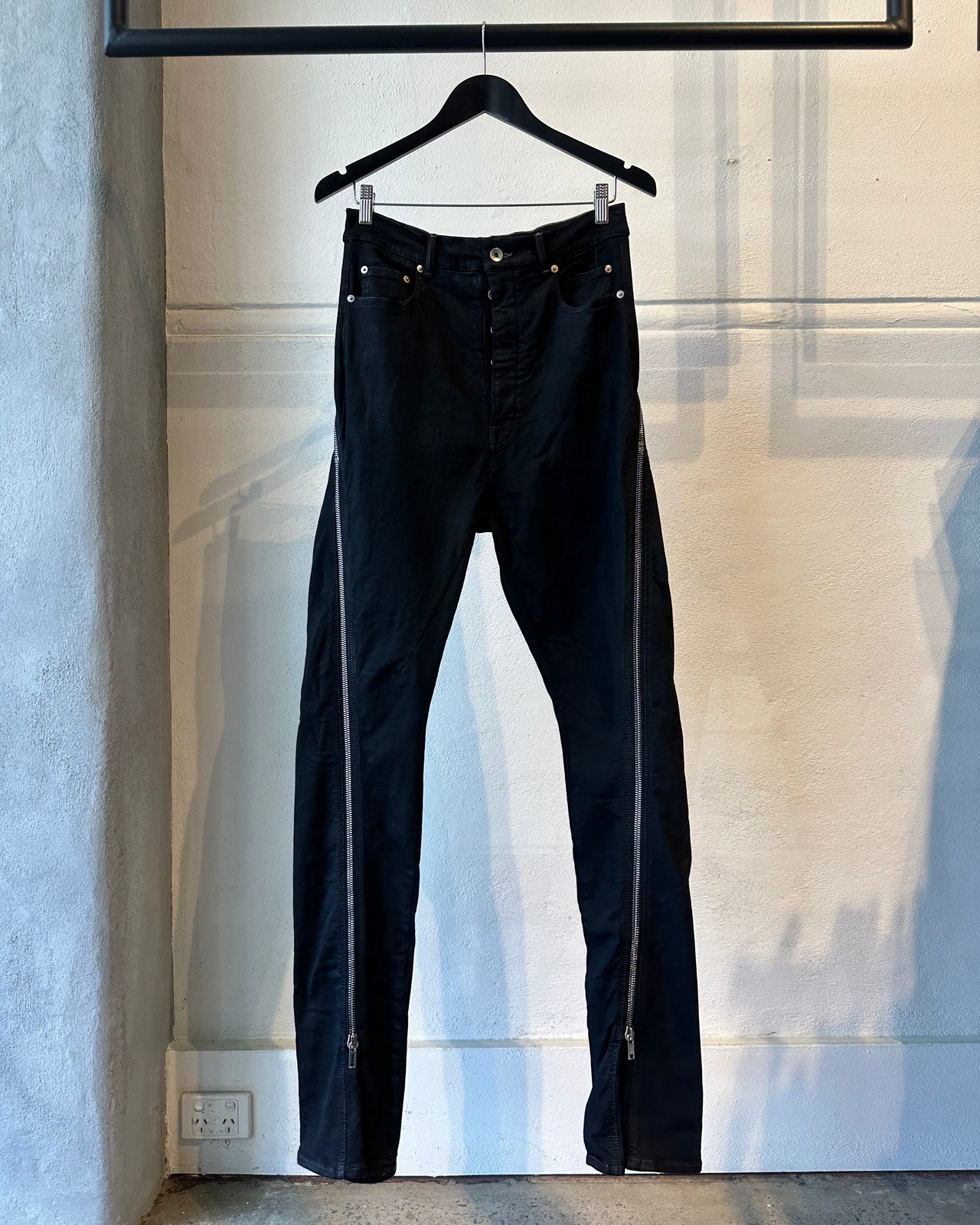 Black Rick Owens jeans hanging on a hanger against a light-colored wall.