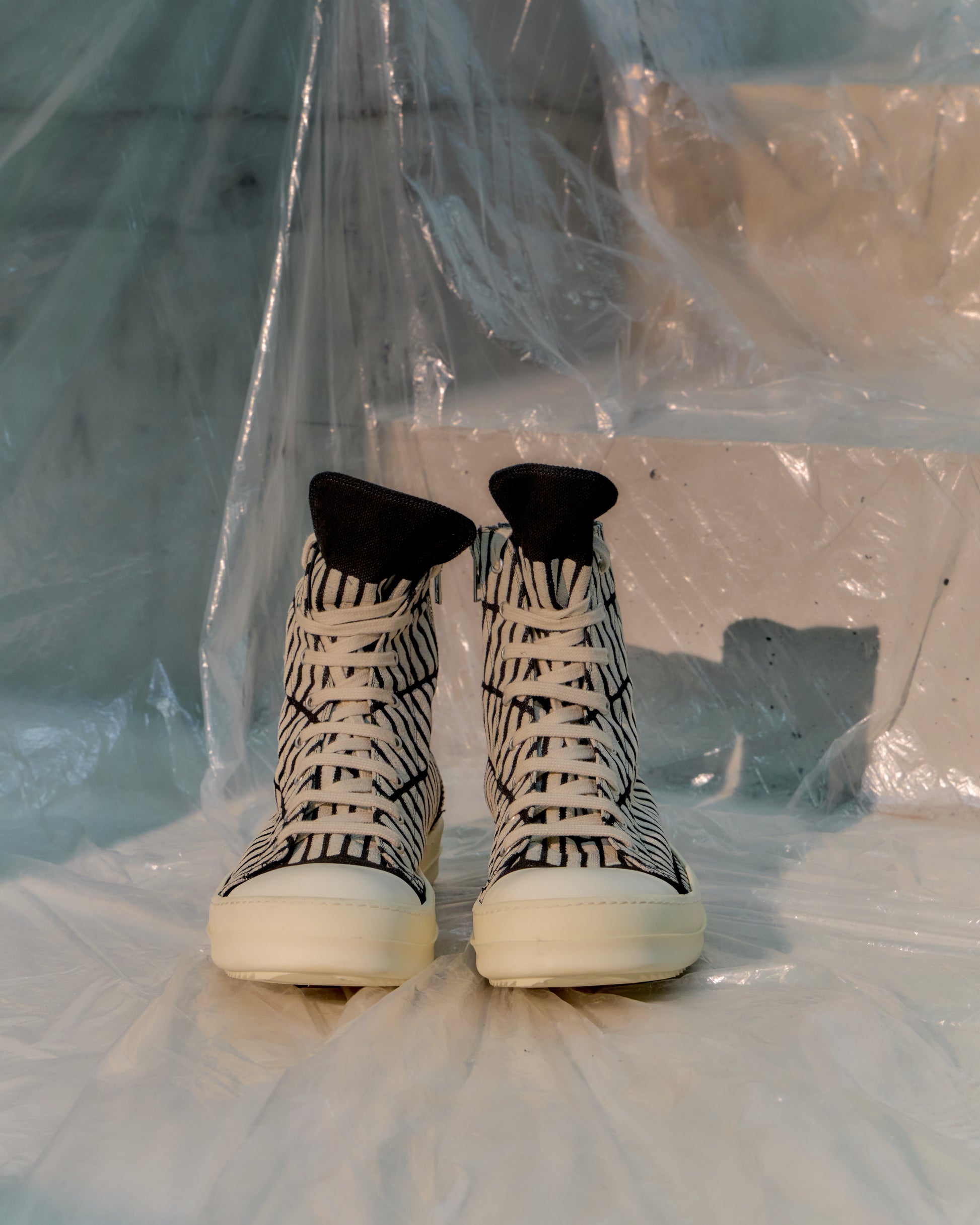 Black and white high-top Rick Owens sneakers on a white surface with a transparent plastic sheet in the background