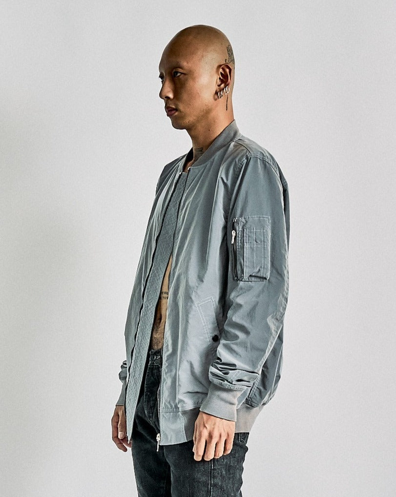 A person with a shaved head and earrings is wearing a loose-fitting, light gray Rick owens bomber jacket over a bare chest. The setting is a neutral background.