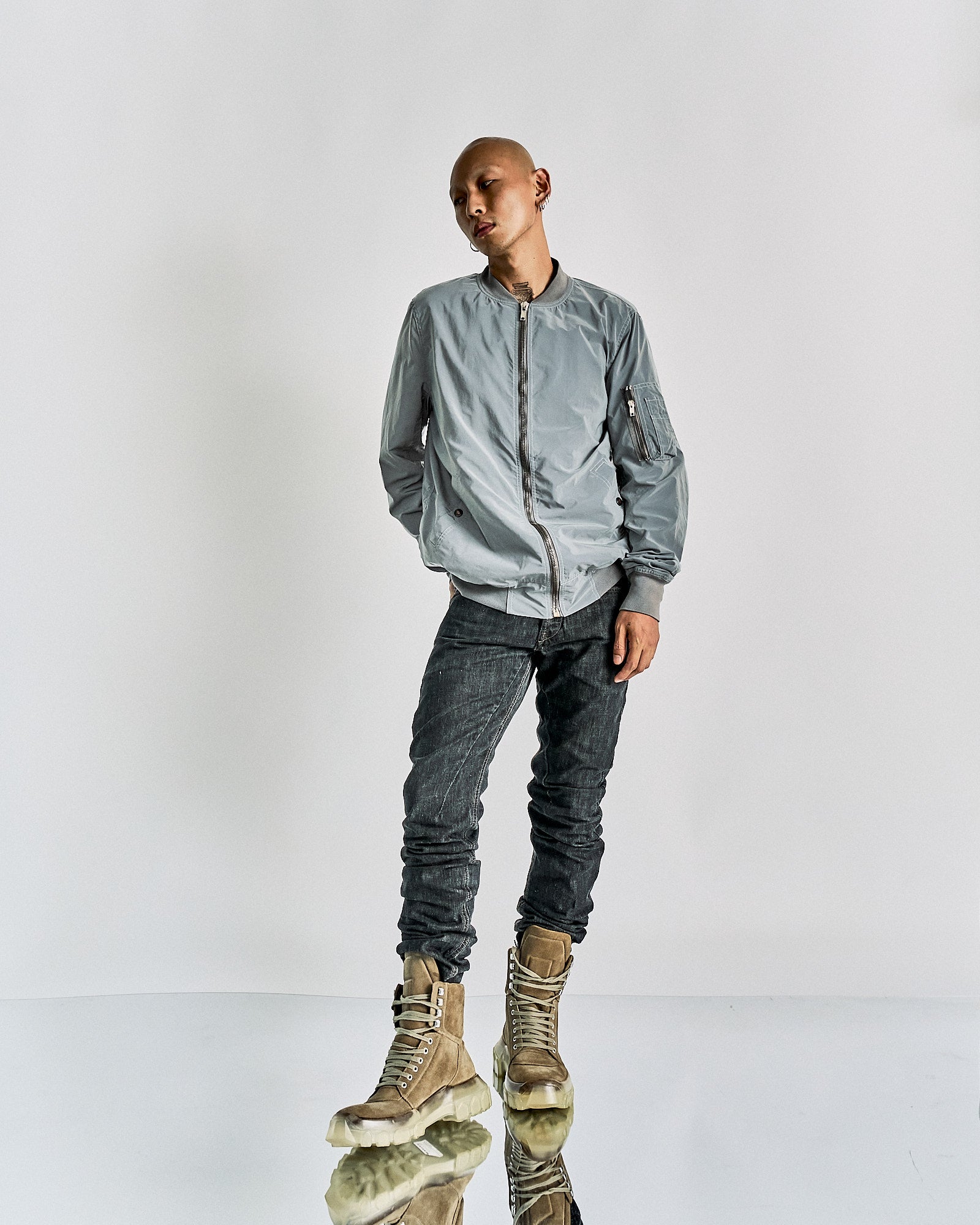 A person in a gray Rick Owens bomber jacket and dark jeans stands confidently with a slight tilt, wearing tan boots.
