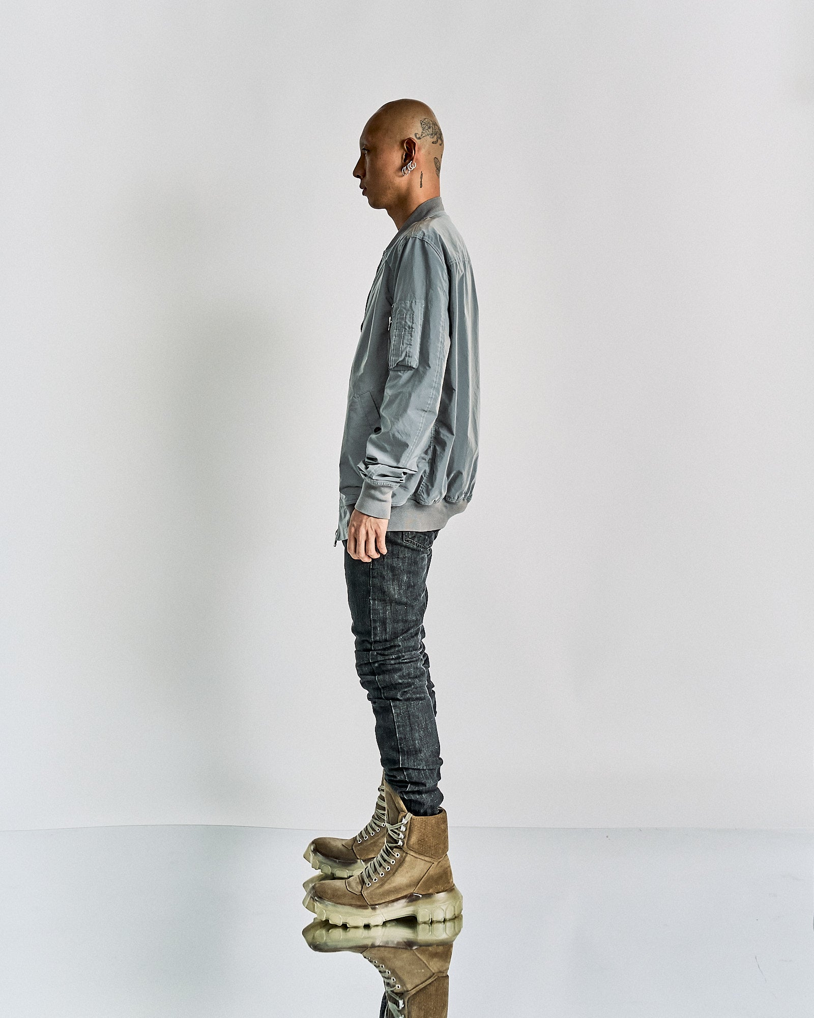 Side profile of a person wearing a light gray Rick Owens jacket, dark jeans, and distinctive tan boots with transparent soles, standing against a plain white background.
