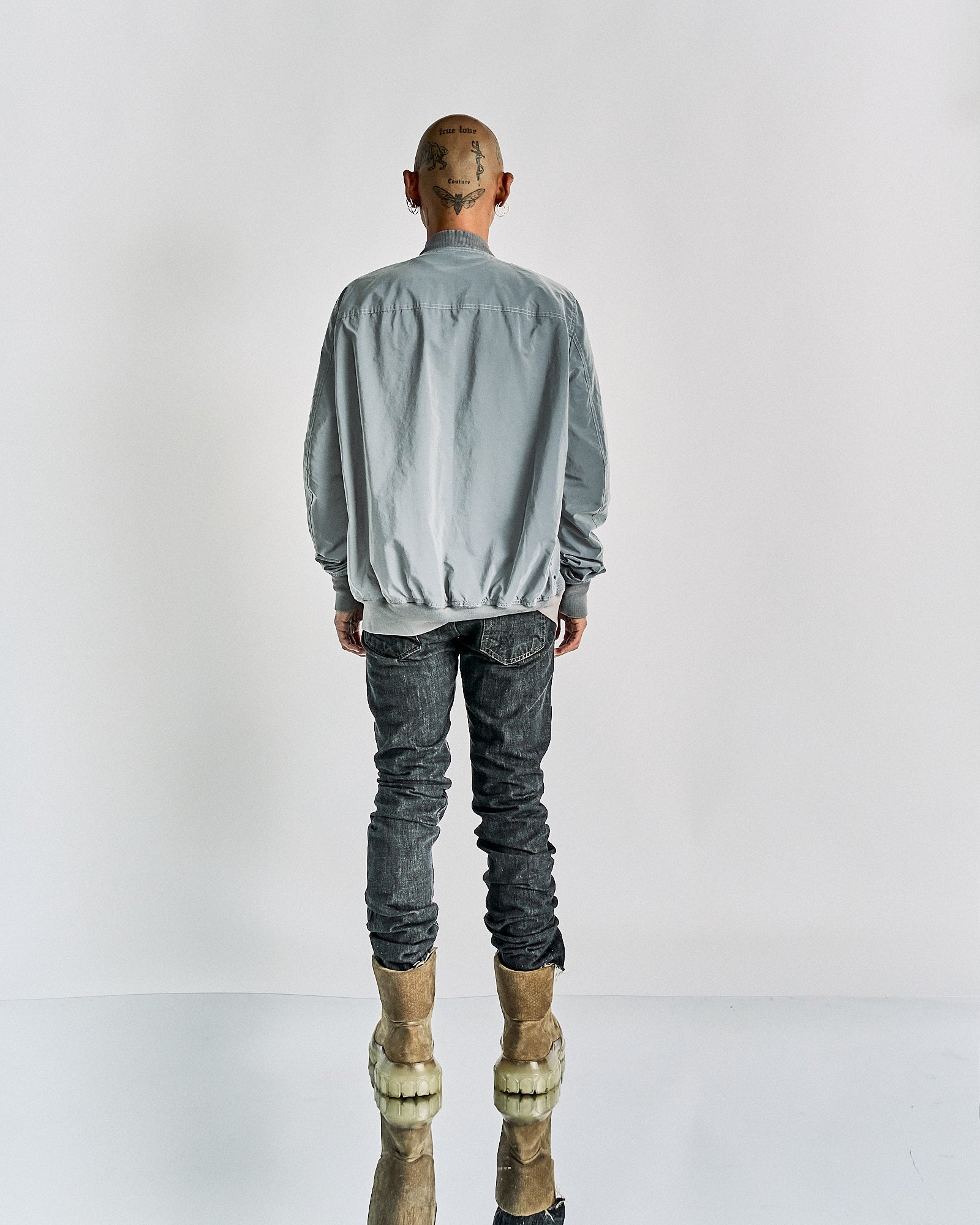 Rear view of a person wearing a light gray Rick Owens jacket, dark jeans, and beige boots with translucent soles. Their shaved head features tattoos. 