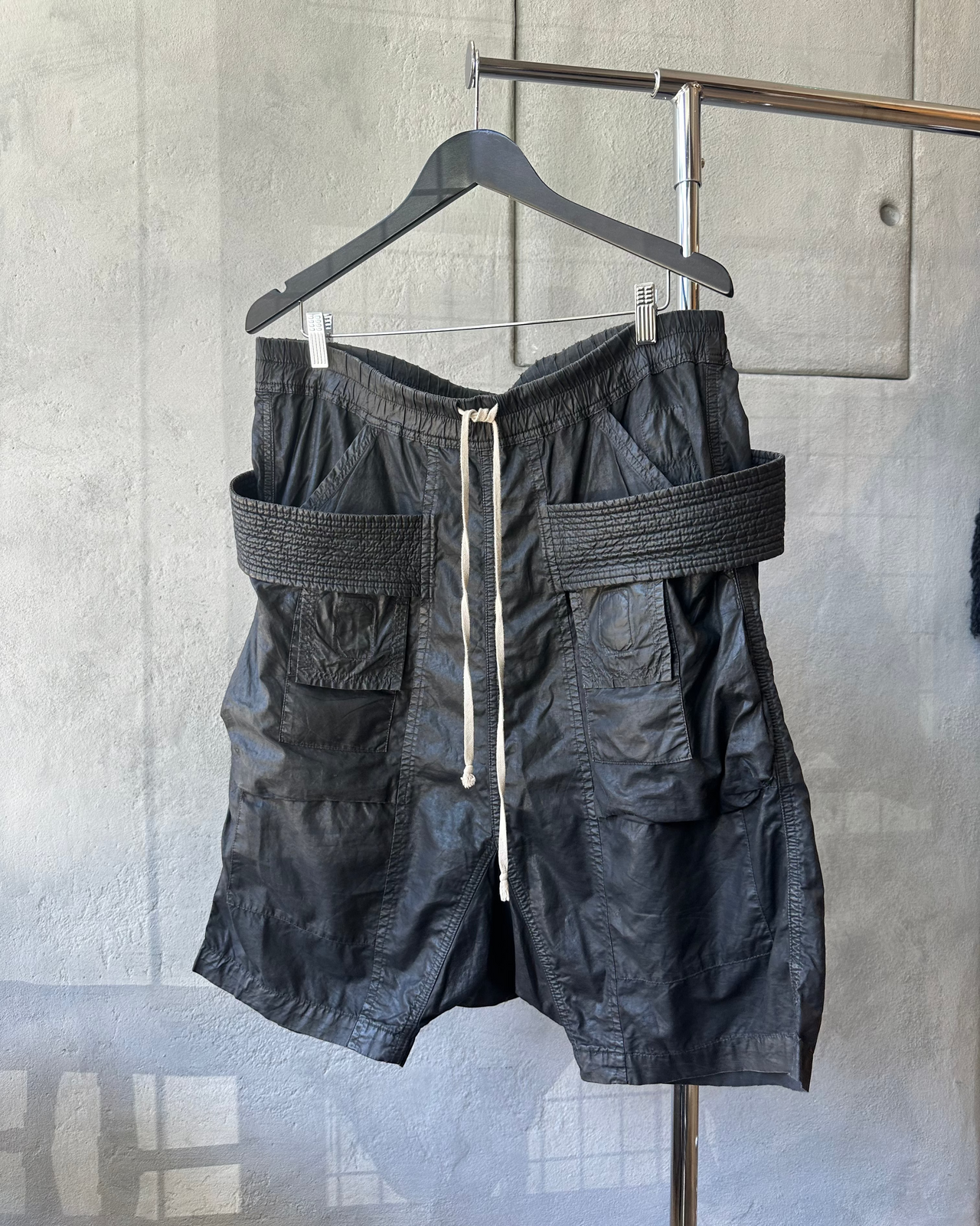 Black Rick Owens shorts with a belt hanging on a hanger against a gray wall.