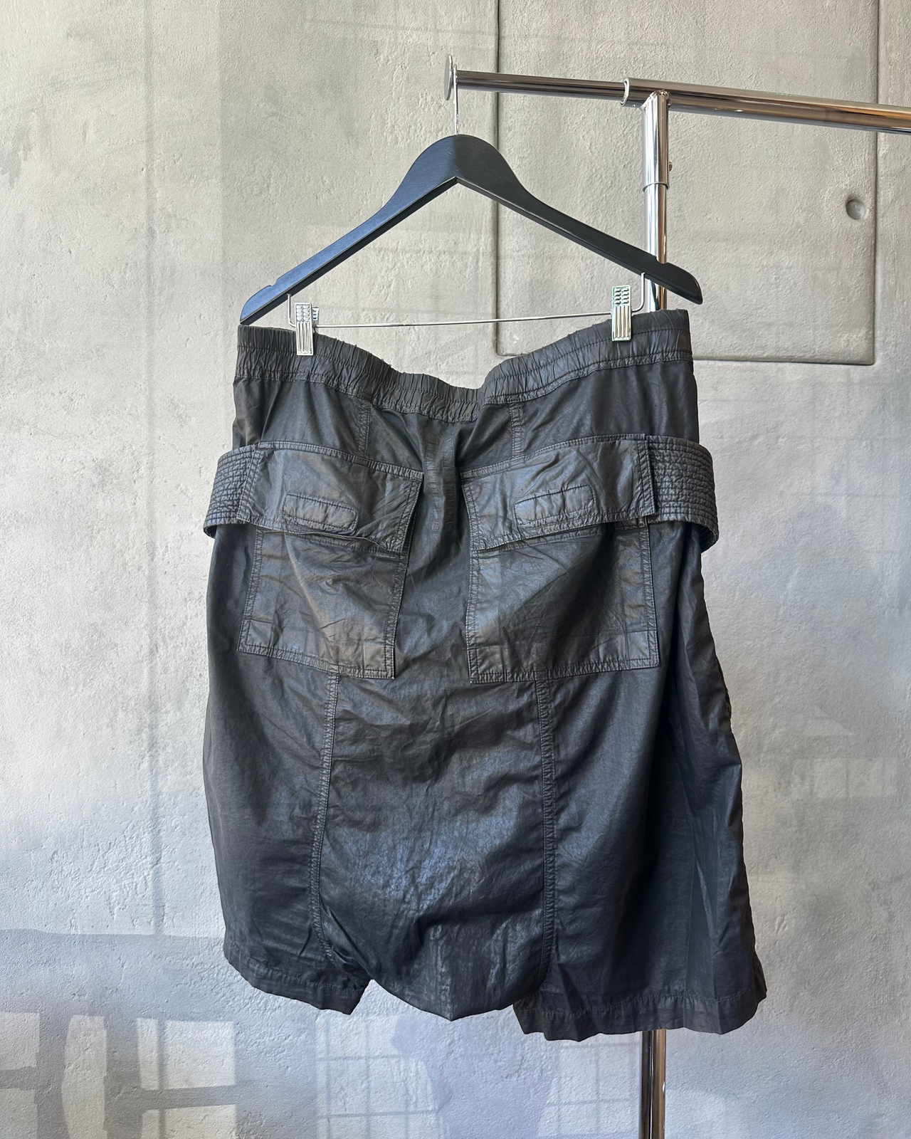Black Rick Owens shorts with a belt hanging on a hanger against a gray wall.
