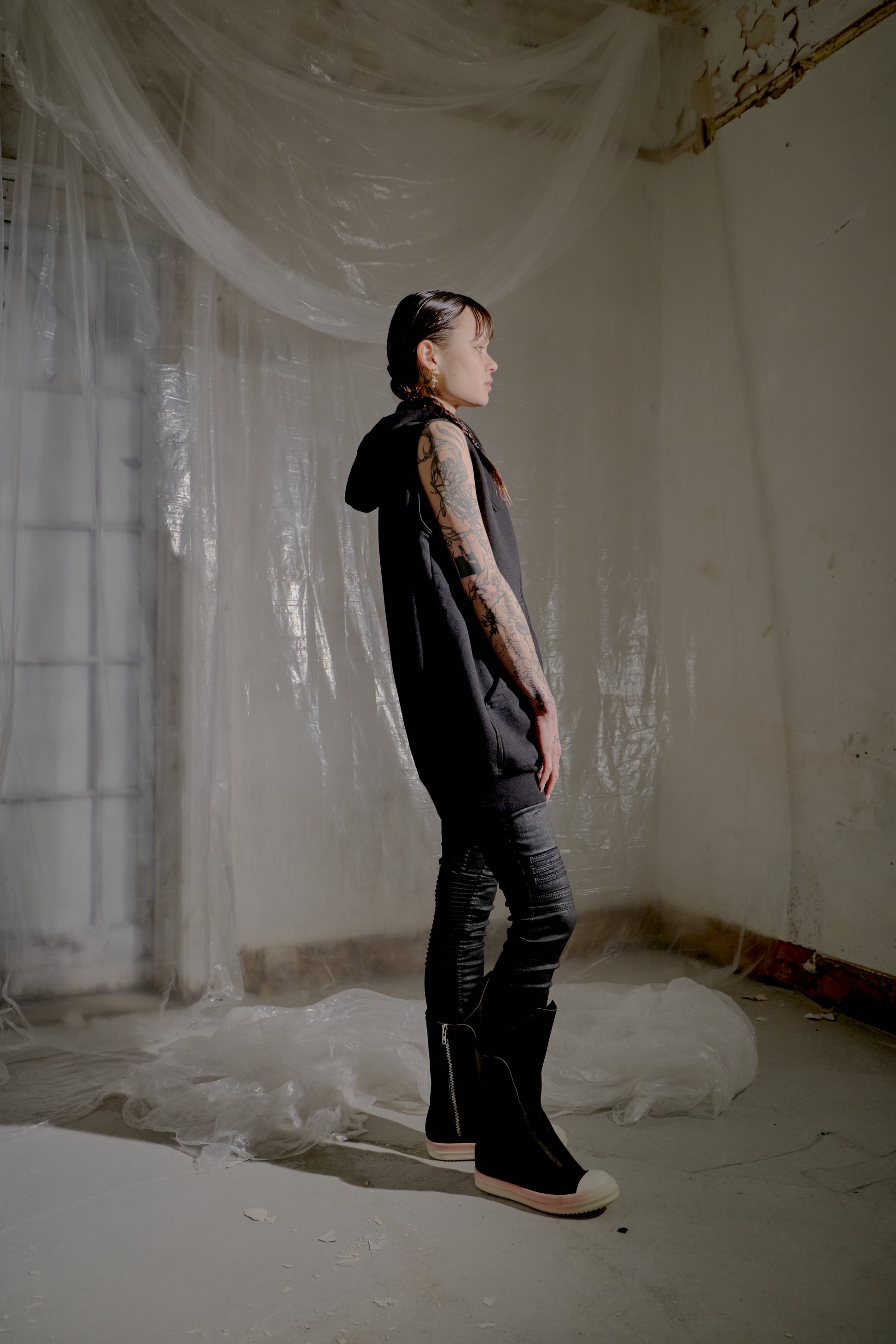 Person standing in a room with white plastic sheeting and a window in the background wearing a black Rick Owens sleeveless jacket.