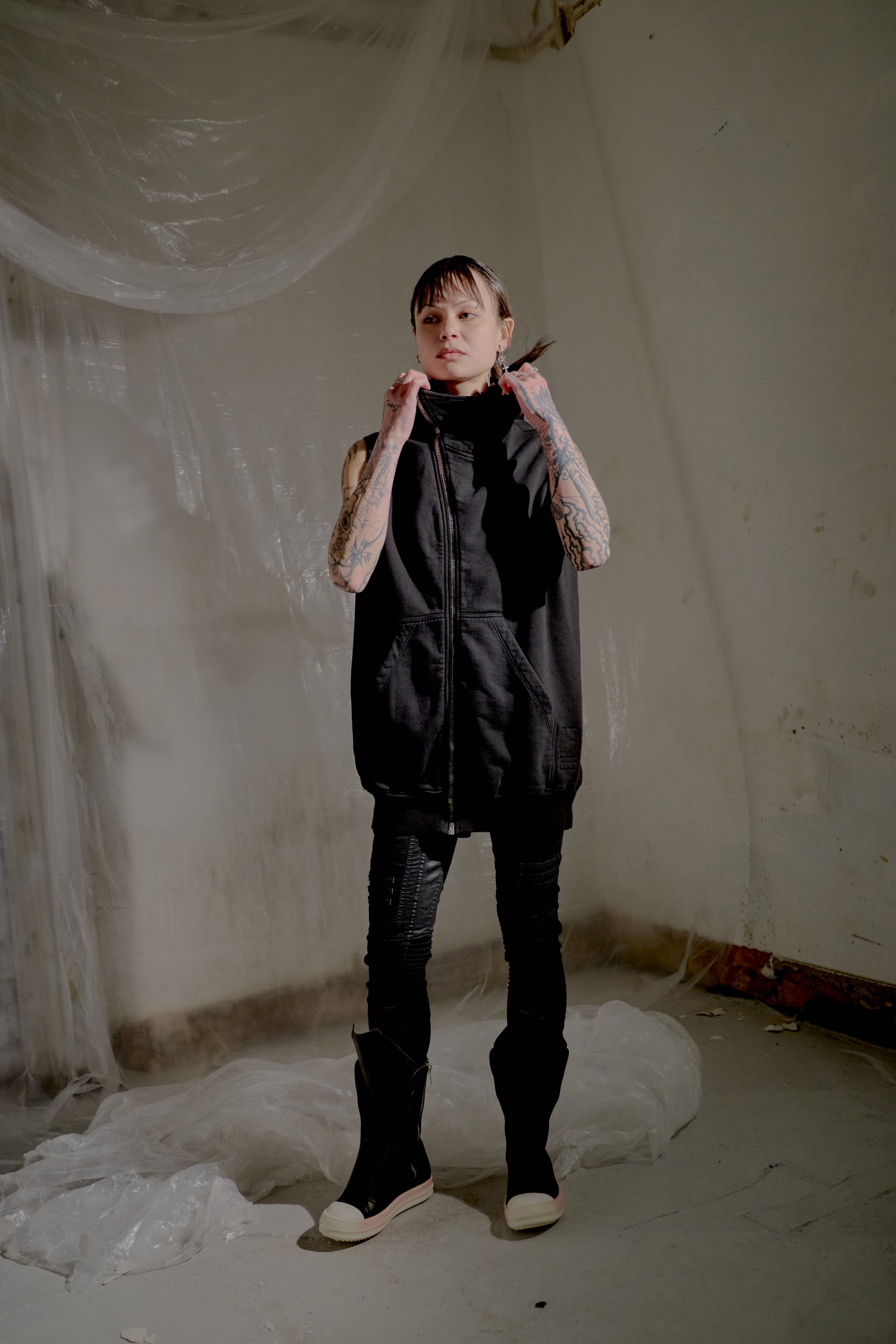 Person wearing a black sleeveless Rick Owens jacket and pants in a minimalistic setting