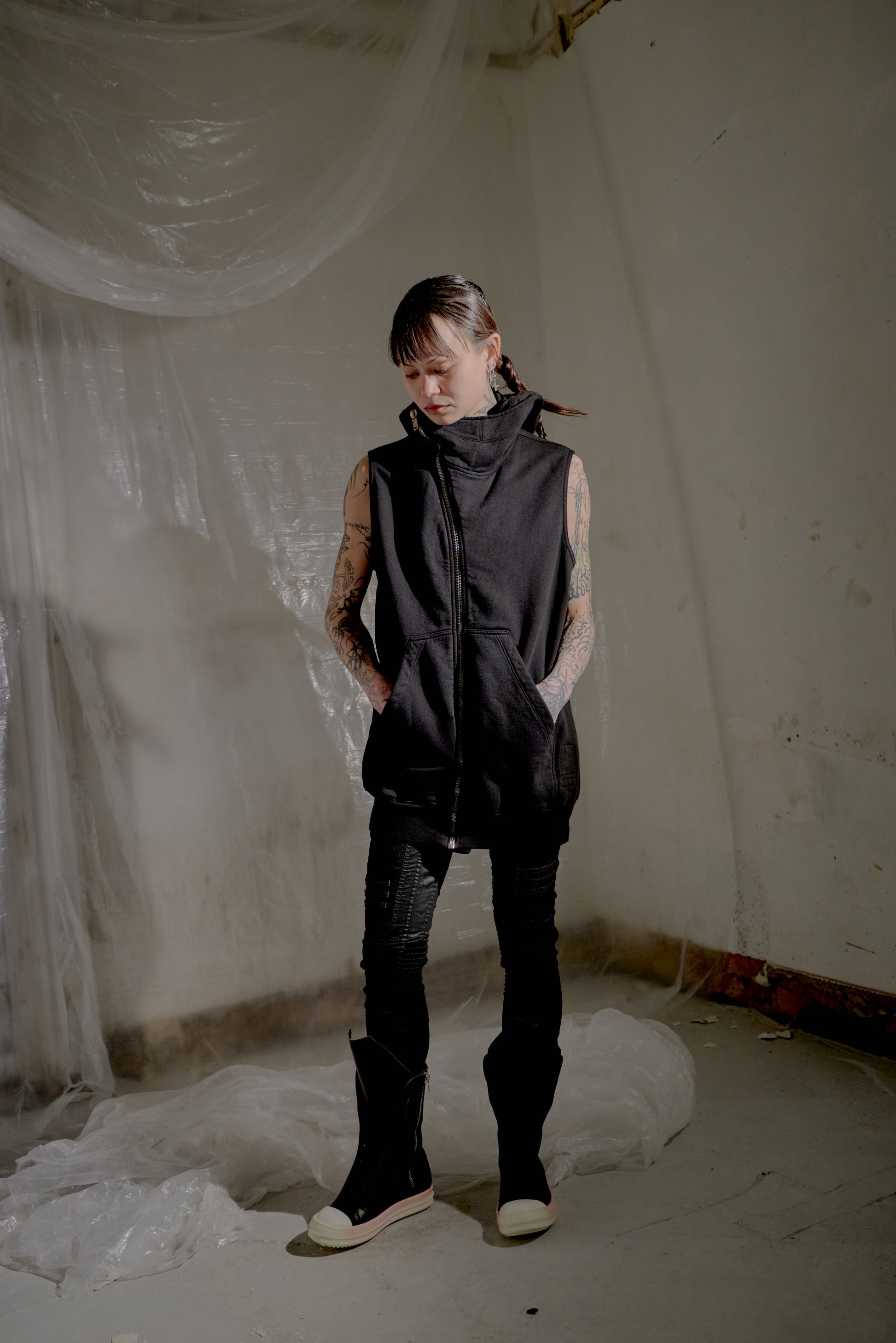 Person wearing a black sleeveless Rick Owens jacket and pants in a minimalistic room with white walls and floor.