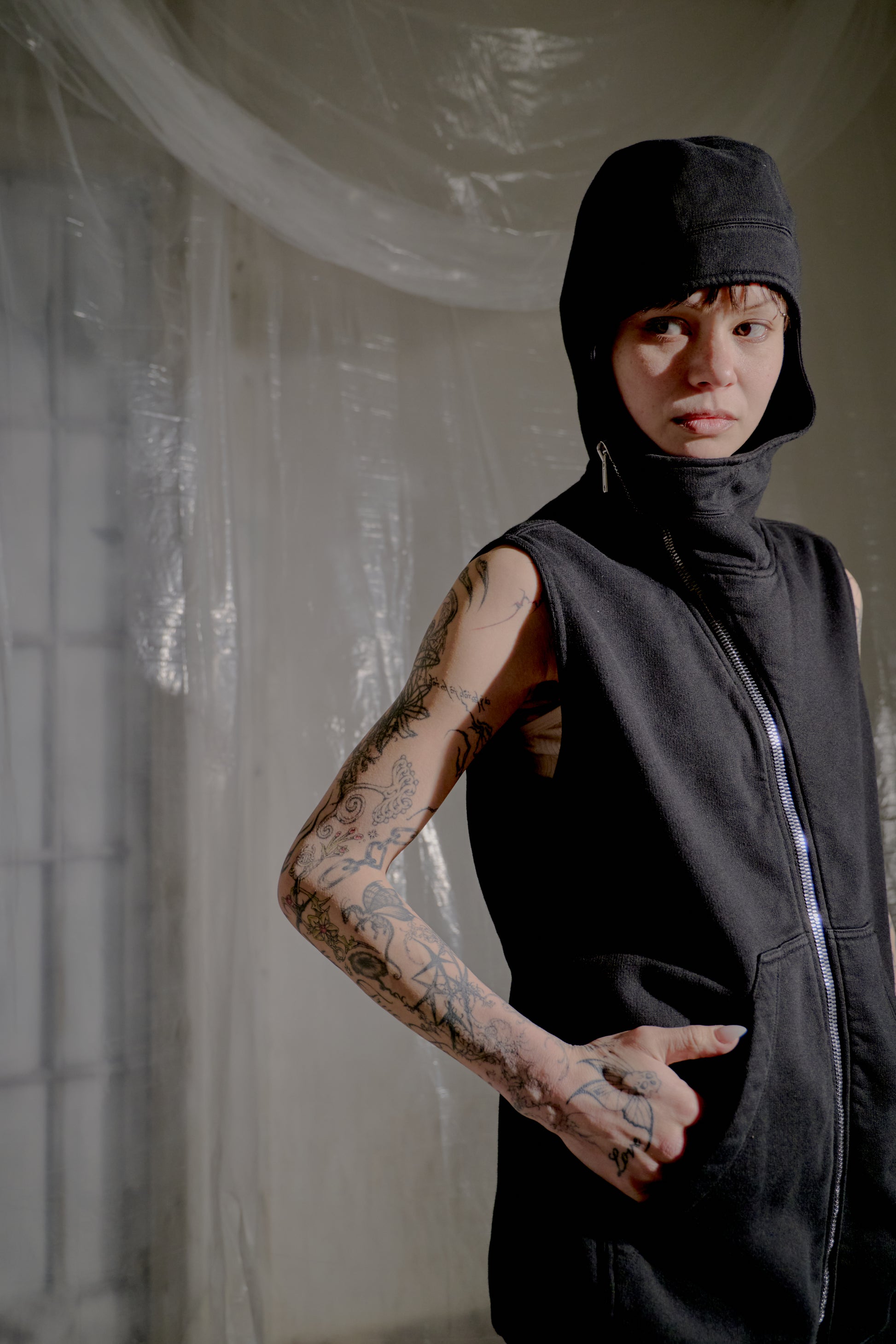 Person wearing a black sleeveless Rick Owens jacket and cap in a minimalistic setting