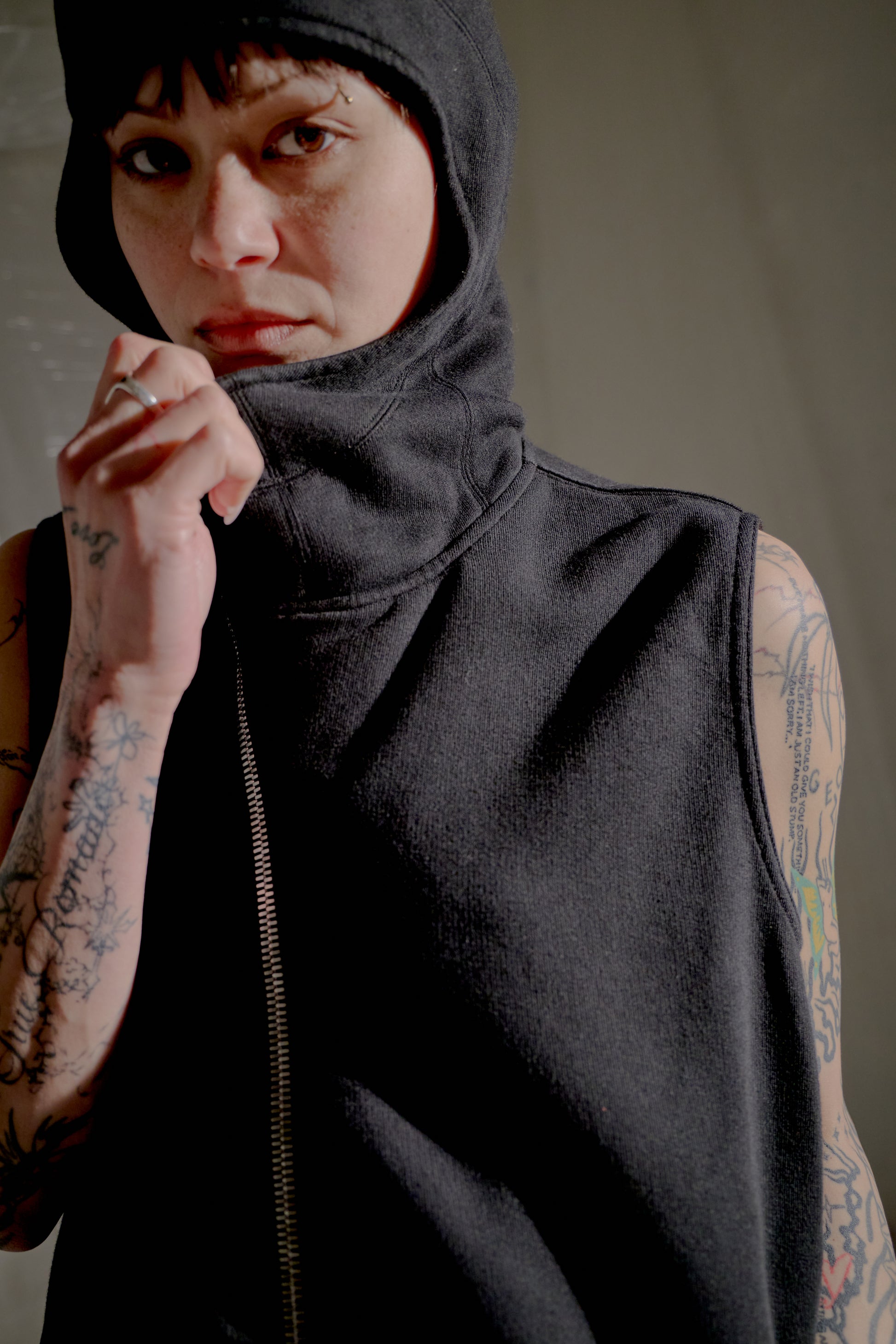 Person wearing a black sleeveless Rick Owens jacket with a hood against a neutral background