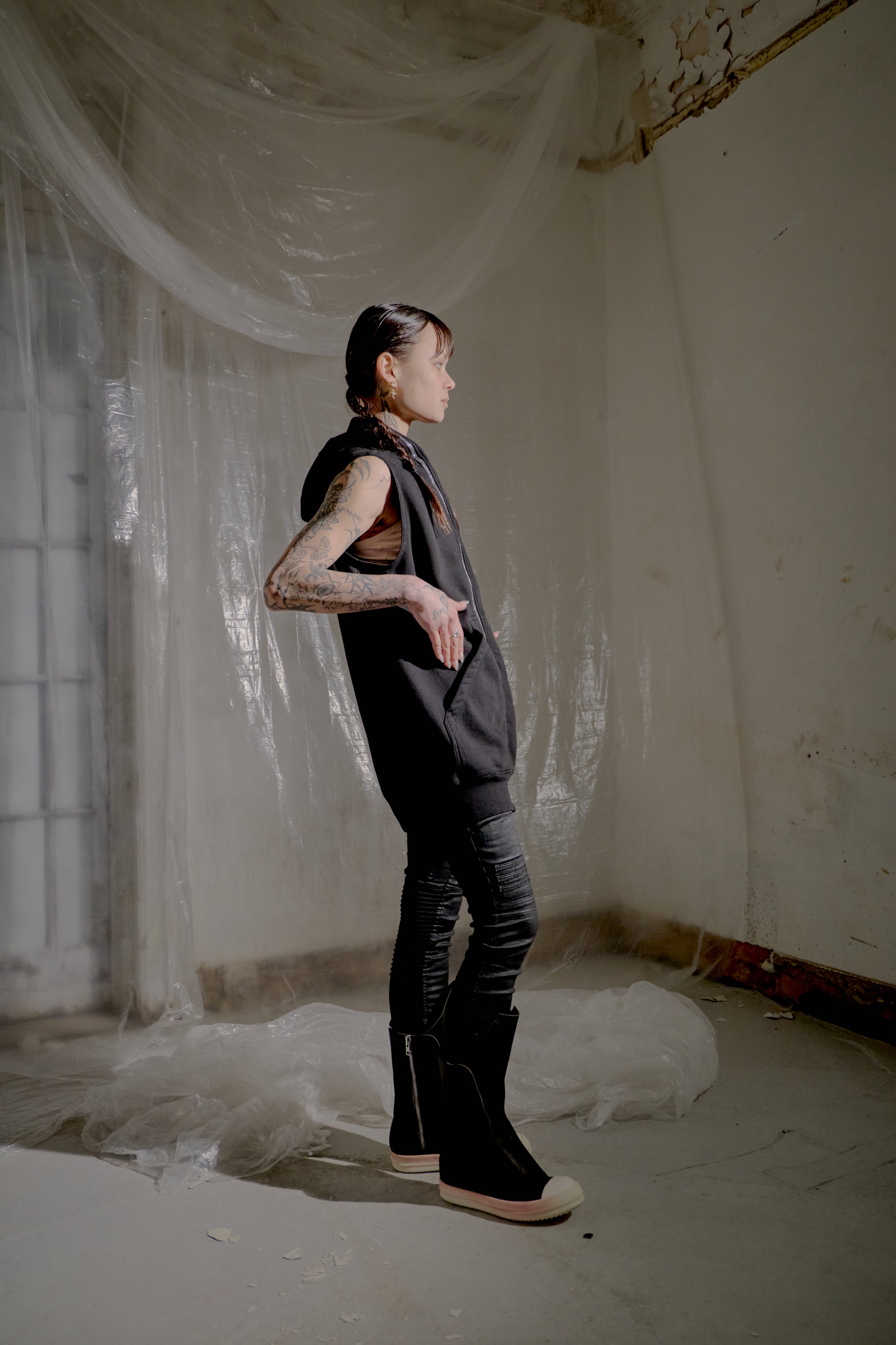 Person standing in a room with white curtains and peeling paint on the walls wearing a black Rick Owens sleeveless jacket. 