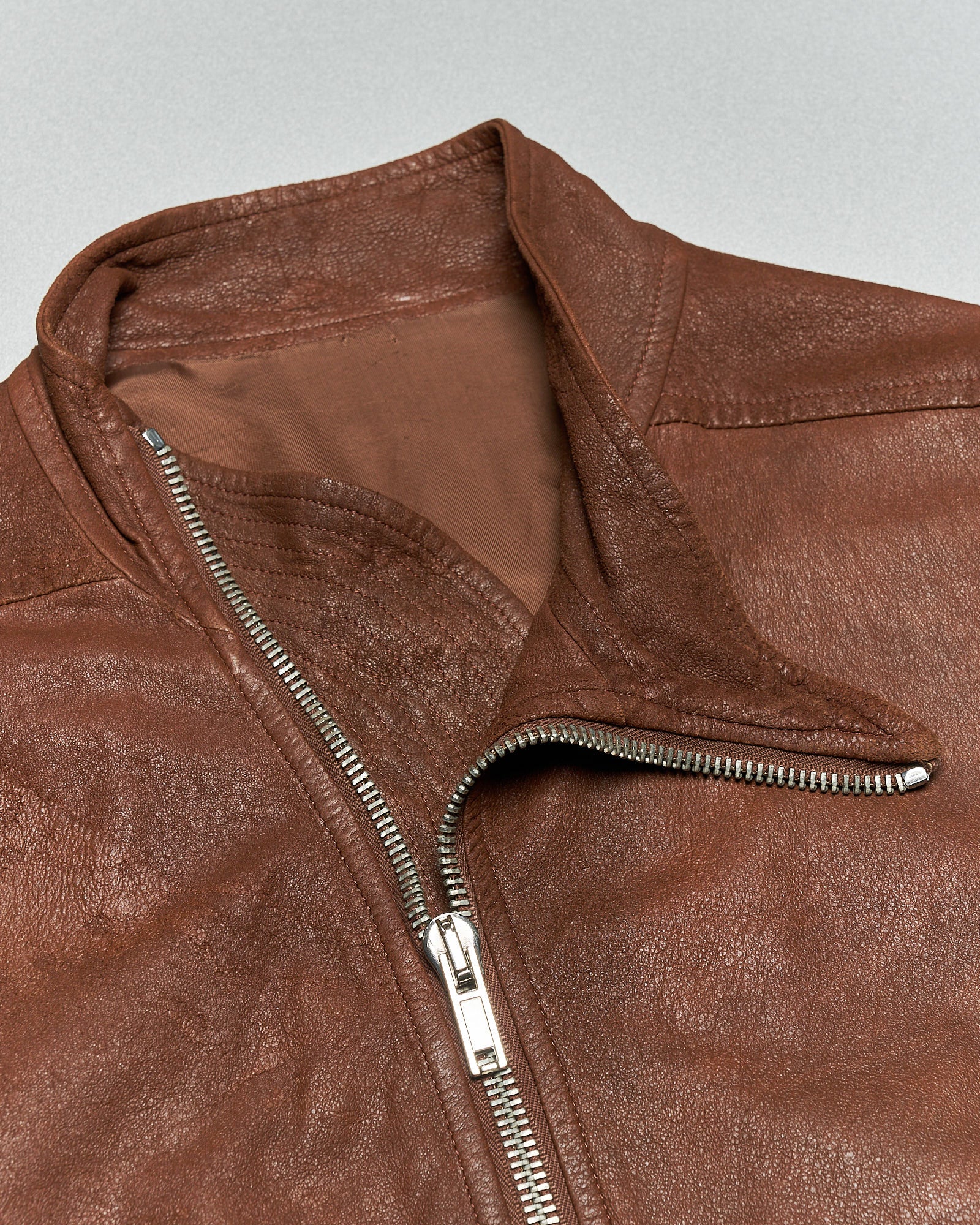 Close-up of a brown leather Rick Owens jacket showing a diagonal silver zipper and raised collar. The texture appears rugged and classic, evoking a vintage feel.