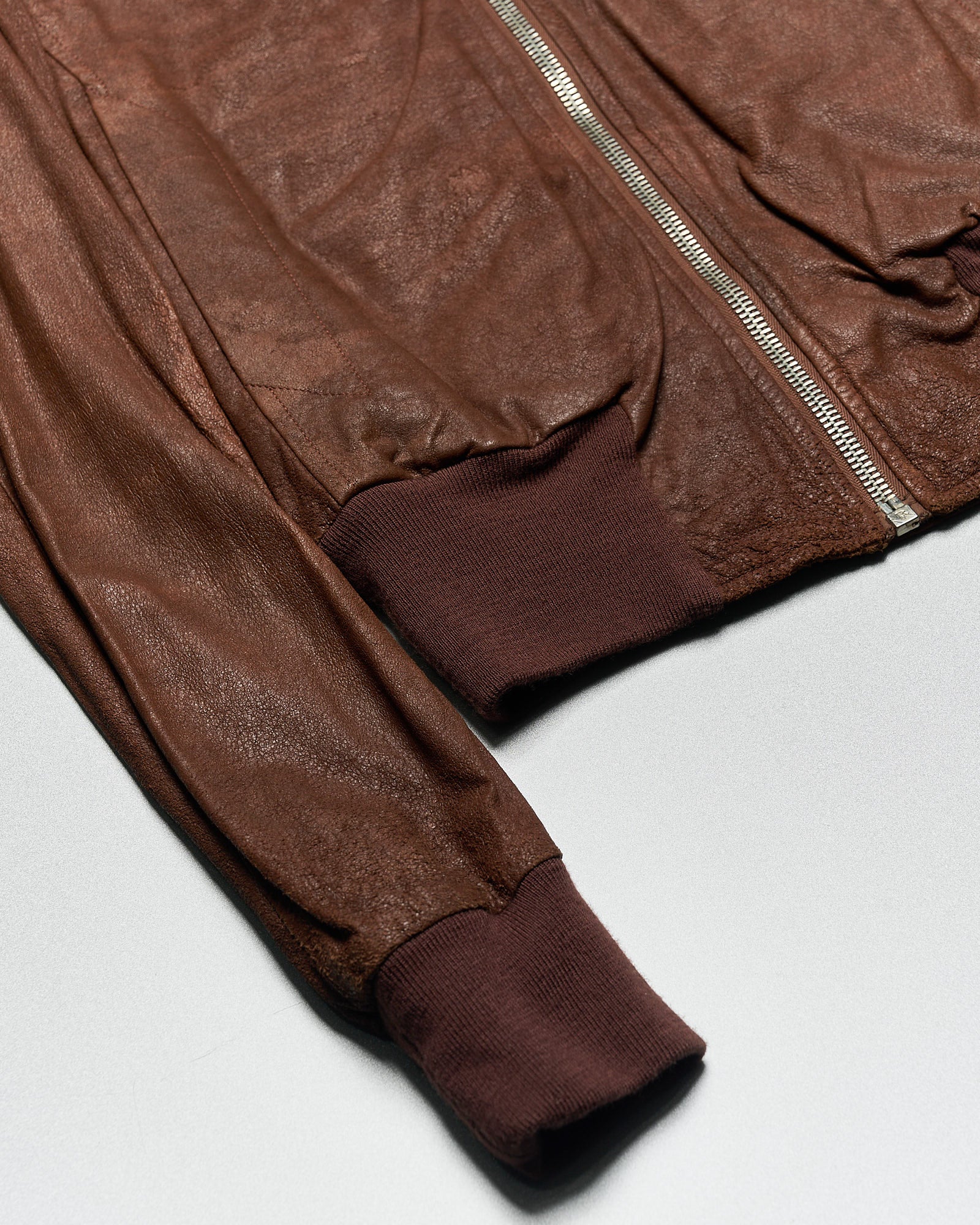 Close-up of a brown leather Rick Owens jacket with a visible silver zipper. The cuffs and waistband are ribbed